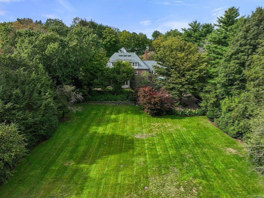 This aerial perspective captures a large, well-manicured lawn leading up to a stately multi-story residence nestled among dense, mature trees. The home features a classic architectural style with a dark shingled roof, multiple dormer windows, and a brick facade. The lush greenery surrounding the property provides a sense of privacy and seclusion, creating a serene and expansive outdoor setting.