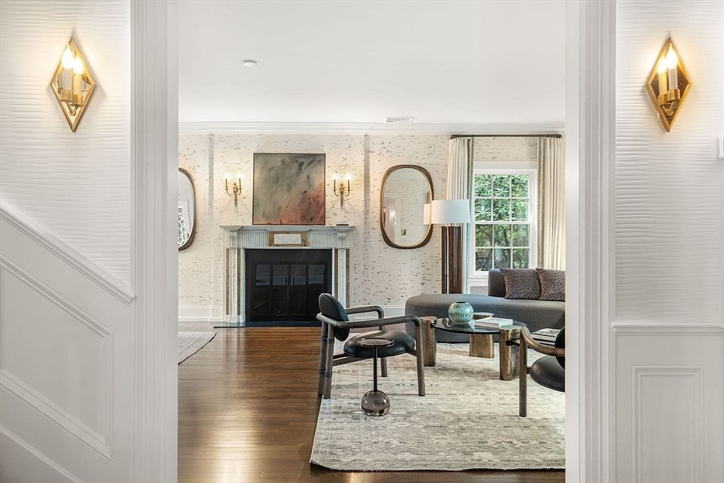 This elegant living room is viewed through a white-trimmed doorway, showcasing a sophisticated design with patterned wallpaper and dark hardwood floors. The space features a central fireplace with a mantel, flanked by wall sconces, and is furnished with a curved sofa and modern armchairs arranged on a textured area rug. The perspective creates a sense of depth, drawing the eye toward the bright window and the refined, contemporary decor.