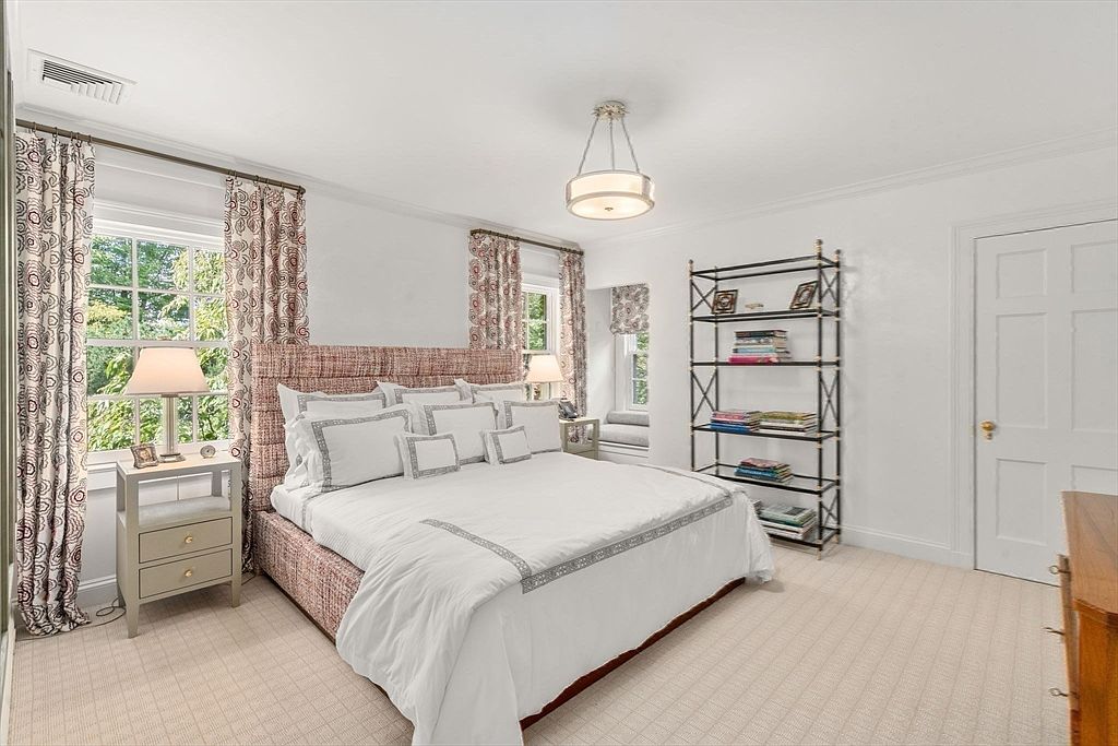 This bright and airy bedroom features a large bed with a textured, patterned headboard and crisp white bedding. The room is accented by patterned curtains framing the windows, a sleek metal shelving unit, and a bedside table with a lamp. The neutral color palette and soft lighting create a serene and inviting atmosphere.