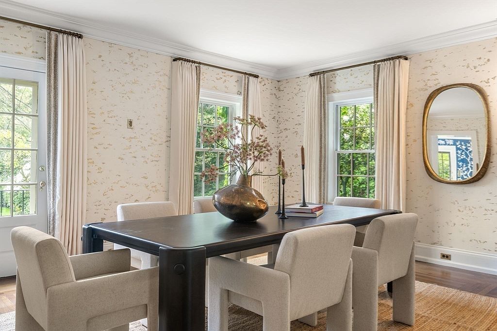 This elegant dining room features a dark, substantial dining table surrounded by six upholstered armchairs in a neutral tone. The walls are adorned with a textured, light-colored wallpaper, and the space is illuminated by natural light streaming through multiple windows dressed in floor-length cream curtains. A decorative vase with branches serves as a centerpiece, while an oval mirror on the wall adds a sophisticated touch to the refined, transitional aesthetic.