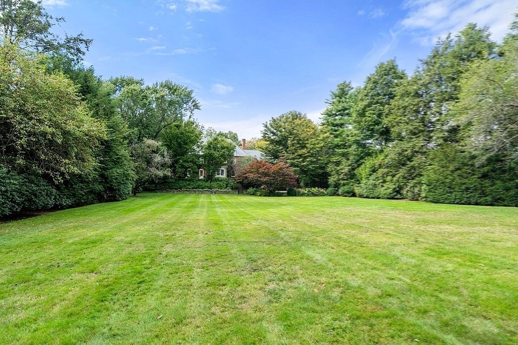 This image captures a expansive, well-manicured lawn that stretches toward a secluded residential home nestled among mature, dense trees. The perspective is from the center of the yard, looking toward the rear of the property, which is framed by lush greenery and a stone wall. The scene conveys a sense of privacy and tranquility, highlighting the generous outdoor space available for recreation or landscaping.