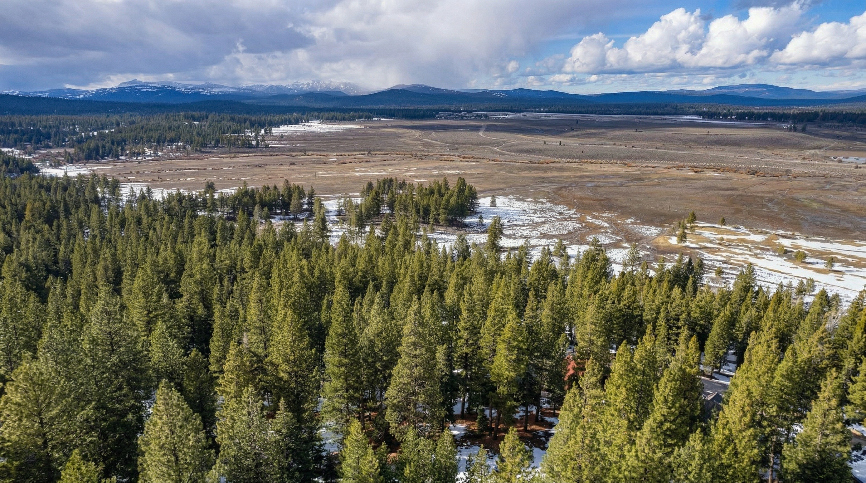 This is a high-altitude aerial view looking down at a heavily forested area nestled in a vast, open valley. The scene captures a mix of dense evergreen trees, snow-dusted ground, and distant snow-capped mountains under a dramatic, cloudy sky. It provides a sense of seclusion and natural beauty, characteristic of a mountainous or rural property setting.