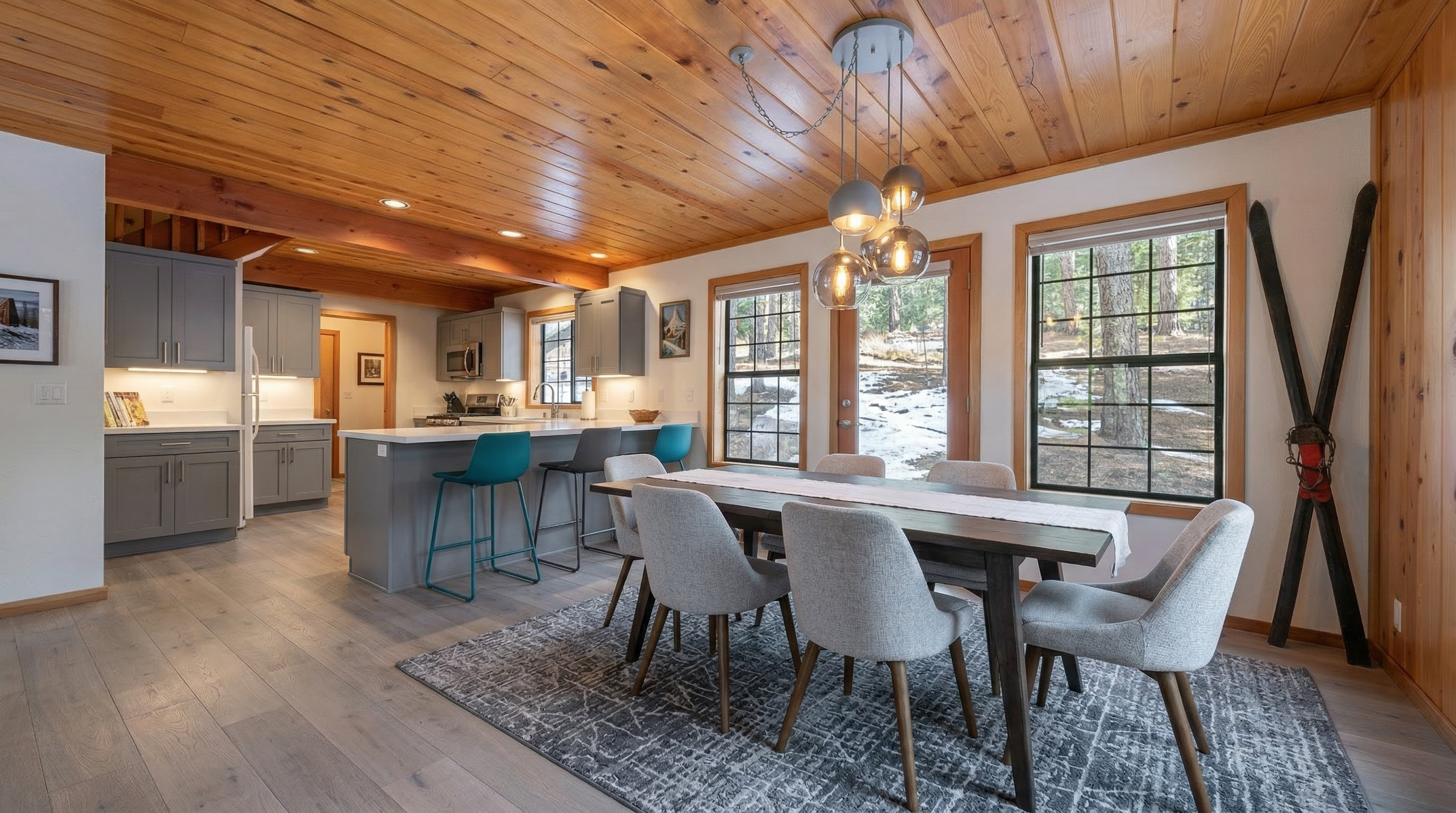 This cozy dining area features warm tongue-and-groove wood paneling on both the walls and ceiling, creating a rustic cabin aesthetic. The room is anchored by a dark wood dining set centered on an intricate area rug, illuminated by a classic pendant chandelier, and is complemented by decorative vintage-style skis leaned against the wall.