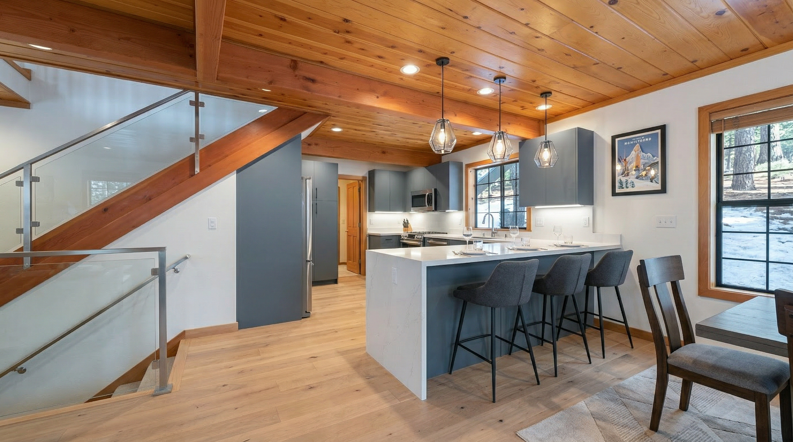 This kitchen area features a rustic cabin aesthetic with natural wood cabinetry and matching wood-paneled ceilings. A breakfast bar is equipped with green stools, providing an inviting space for casual dining. The room flows into an adjoining area with stairs, and a large window lets in ample natural light, highlighting the cozy, mountain-lodge atmosphere.