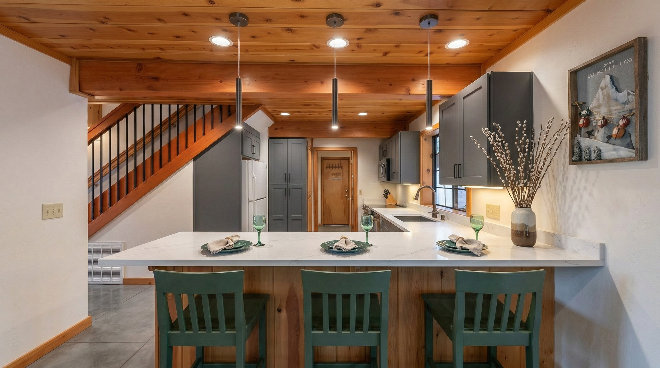 This rustic kitchen features warm natural wood cabinetry, exposed wood ceiling beams, and a charming breakfast bar with stylish teal chairs. The open-concept design transitions seamlessly into the living space, highlighted by cozy cabin-inspired decor and a view of an adjacent staircase. It offers a warm, inviting atmosphere perfect for casual dining and social gatherings.