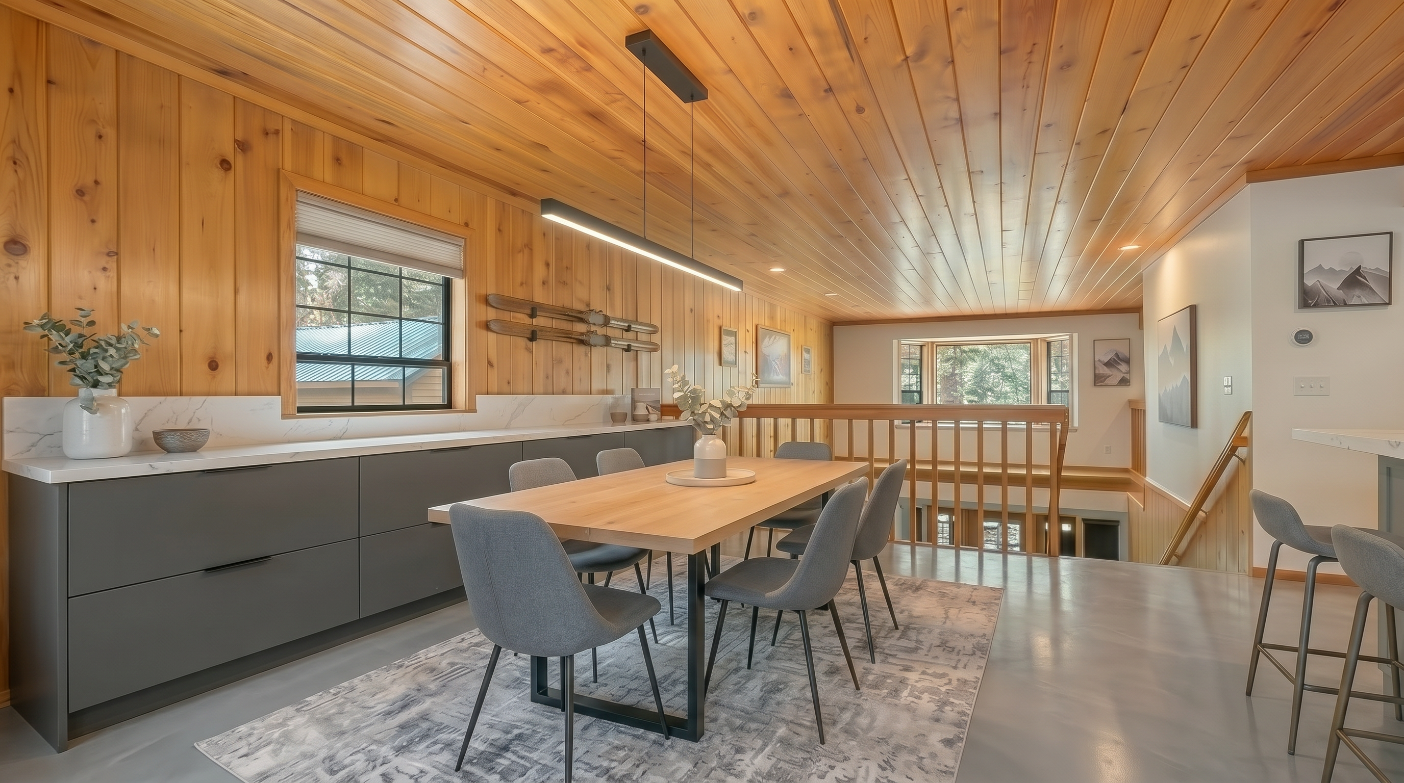 This spacious, open-concept dining room exudes a warm, rustic charm with its extensive wood-paneled walls and ceilings. The area features a large modern dining table, comfortable upholstered chairs, and a sleek kitchen island in the background, creating an inviting space for family gatherings. Highlighting the home's contemporary feel, the room is illuminated by a unique multi-globe pendant light fixture and offers an open layout that seamlessly connects to the kitchen and staircase.