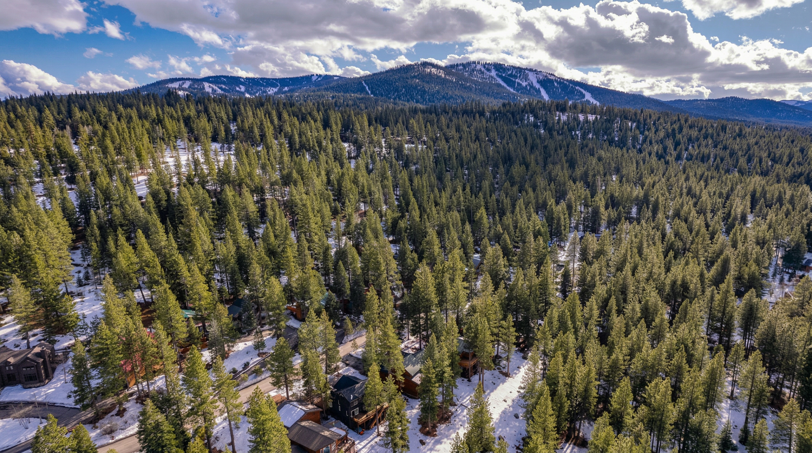 This high-altitude aerial view showcases a residential enclave nestled within a dense forest of evergreen trees. The scene features a mix of multi-story mountain homes characterized by wood siding and steep, snow-shedding rooflines, surrounded by partly snow-covered landscapes. A white directional arrow highlights one of the properties, emphasizing its secluded location framed by scenic, snow-capped mountain peaks under a vibrant, cloudy sky.