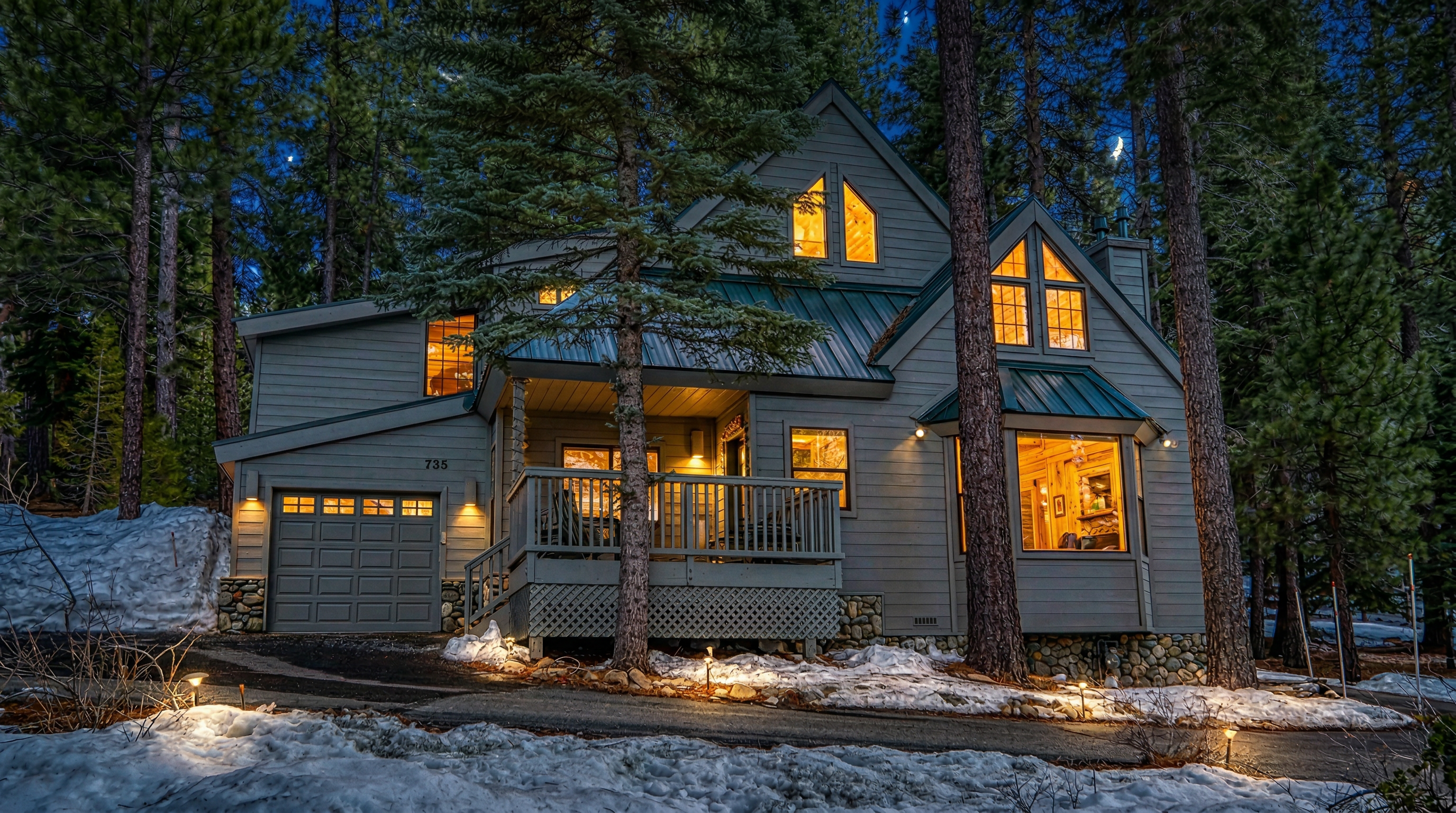 This charming, multi-level mountain home features light gray siding with a contrasting green metal roof, nestled in a lush, snow-covered forest setting. The exterior includes a prominent garage, an inviting front deck with lattice detailing, and large windows that allow for plenty of natural light. The property exudes a cozy, rustic-chic appeal, perfectly suited for a alpine retreat.