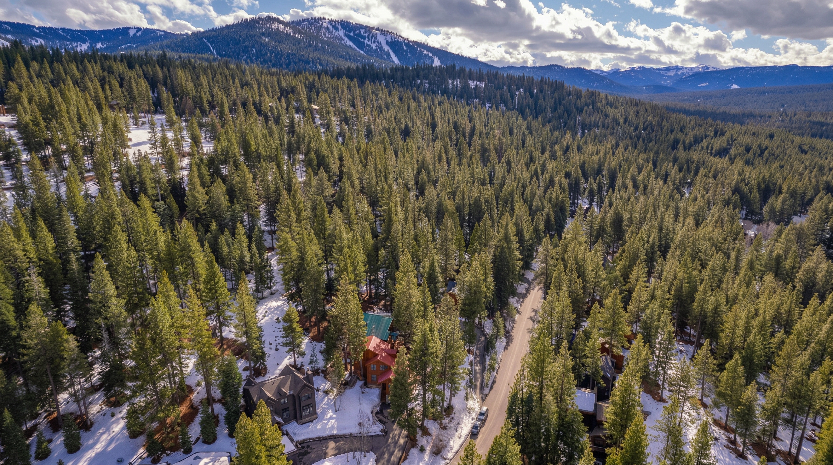 This high-altitude aerial view showcases a picturesque residential setting nestled within a dense, snow-covered pine forest. A winding road weaves through the treeline, providing access to several homes, including a standout property with a prominent red-roofed structure. The backdrop features dramatic mountain ranges under a sky filled with scattered clouds, emphasizing a serene, mountainous environment.