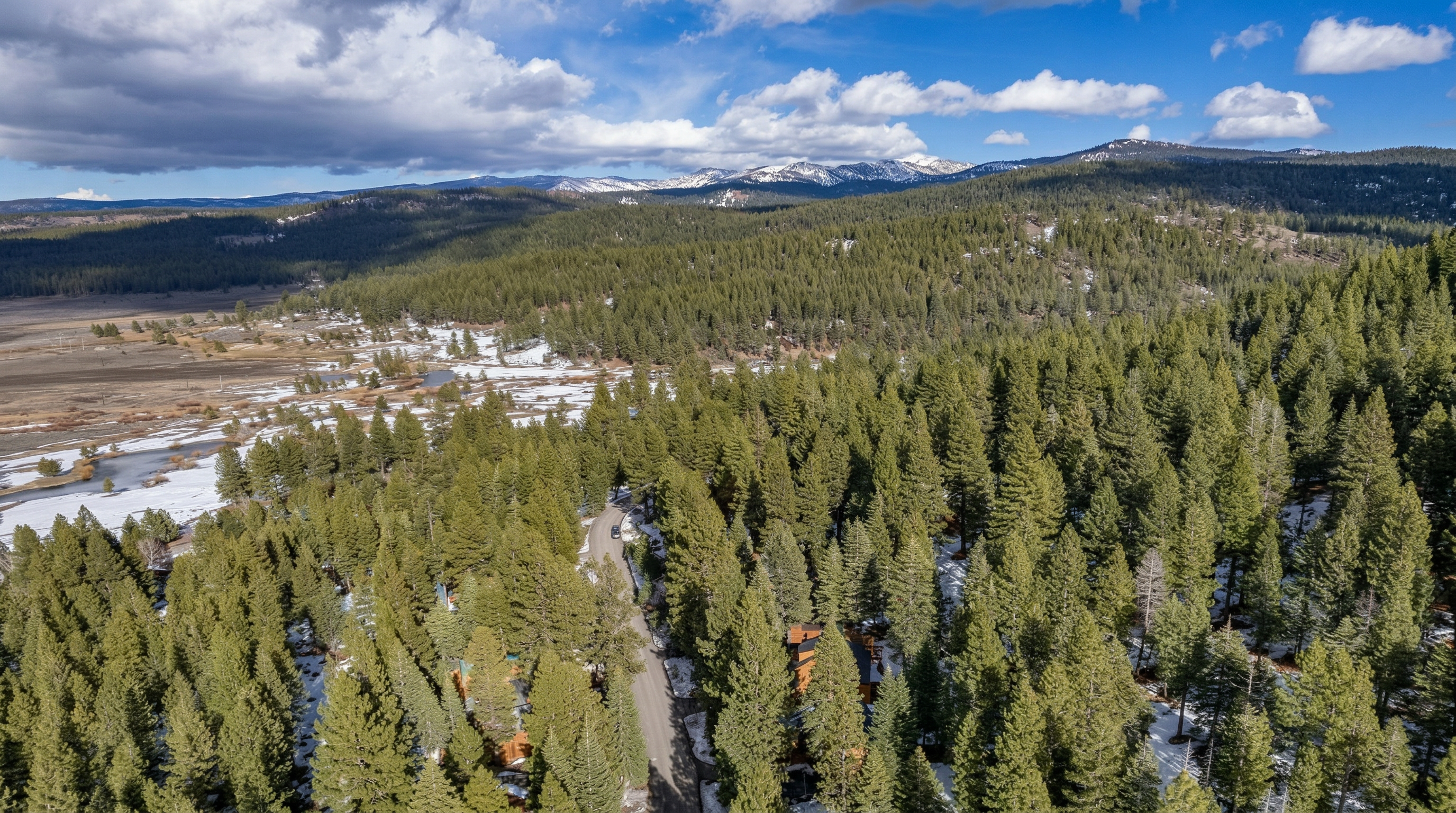 This aerial view captures a rustic wooden cabin nestled within a dense evergreen forest, set against a backdrop of snow-capped mountains. The perspective highlights the property's secluded and natural wilderness setting, with a paved road providing access to the residence. It is an ideal property for those seeking a tranquil retreat surrounded by an expansive alpine landscape.