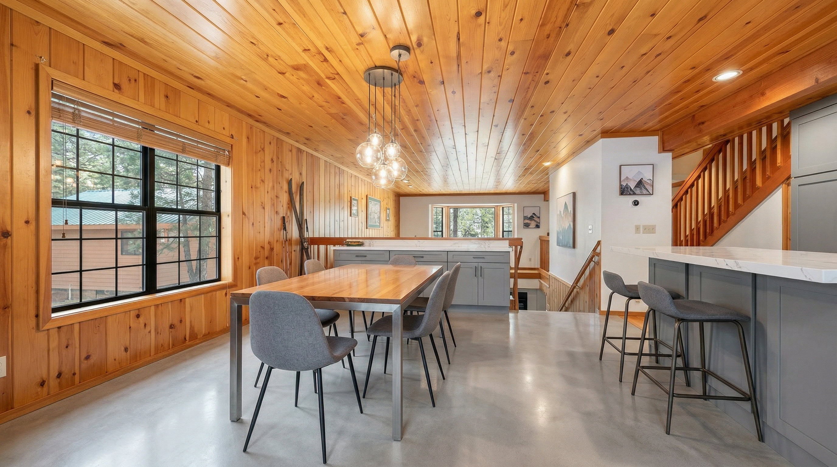 This rustic dining room features warm wood-paneled walls and ceilings, complemented by a central wooden table surrounded by matching chairs. A patterned area rug anchors the space, while a decorative pendant light hangs above the table, creating an inviting atmosphere. The room also showcases architectural interest with an adjacent staircase and views through large windows, embodying a cozy, cabin-like aesthetic.