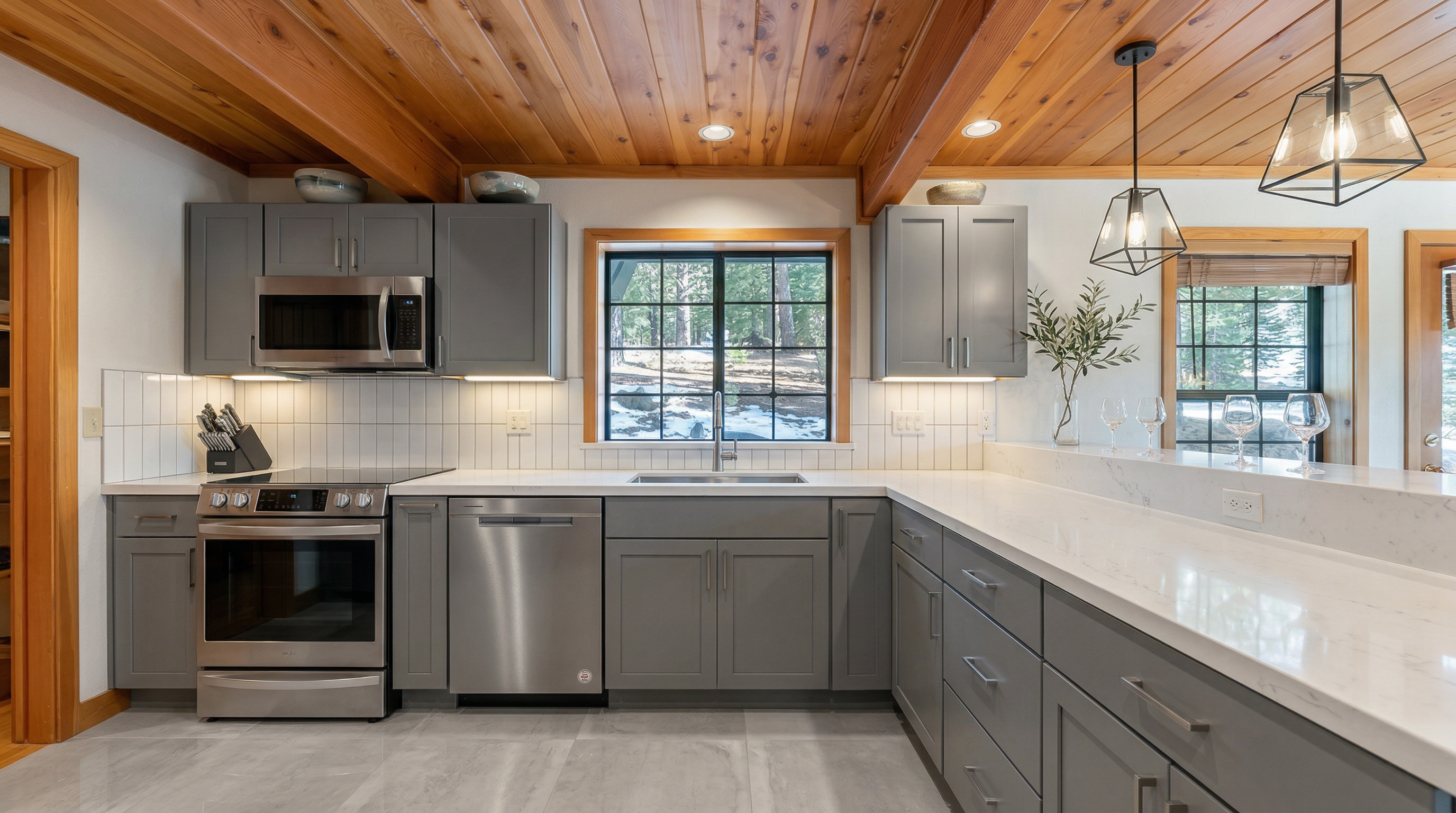 This rustic, cabin-style kitchen features warm natural wood cabinetry, a tongue-and-groove wood ceiling, and rich granite countertops. The space is well-equipped with stainless steel appliances, including a dishwasher and a built-in microwave, while a large window above the sink provides ample natural light and forest views. The combination of wood textures and stone surfaces creates a cozy, inviting atmosphere characteristic of a mountain or vacation home.