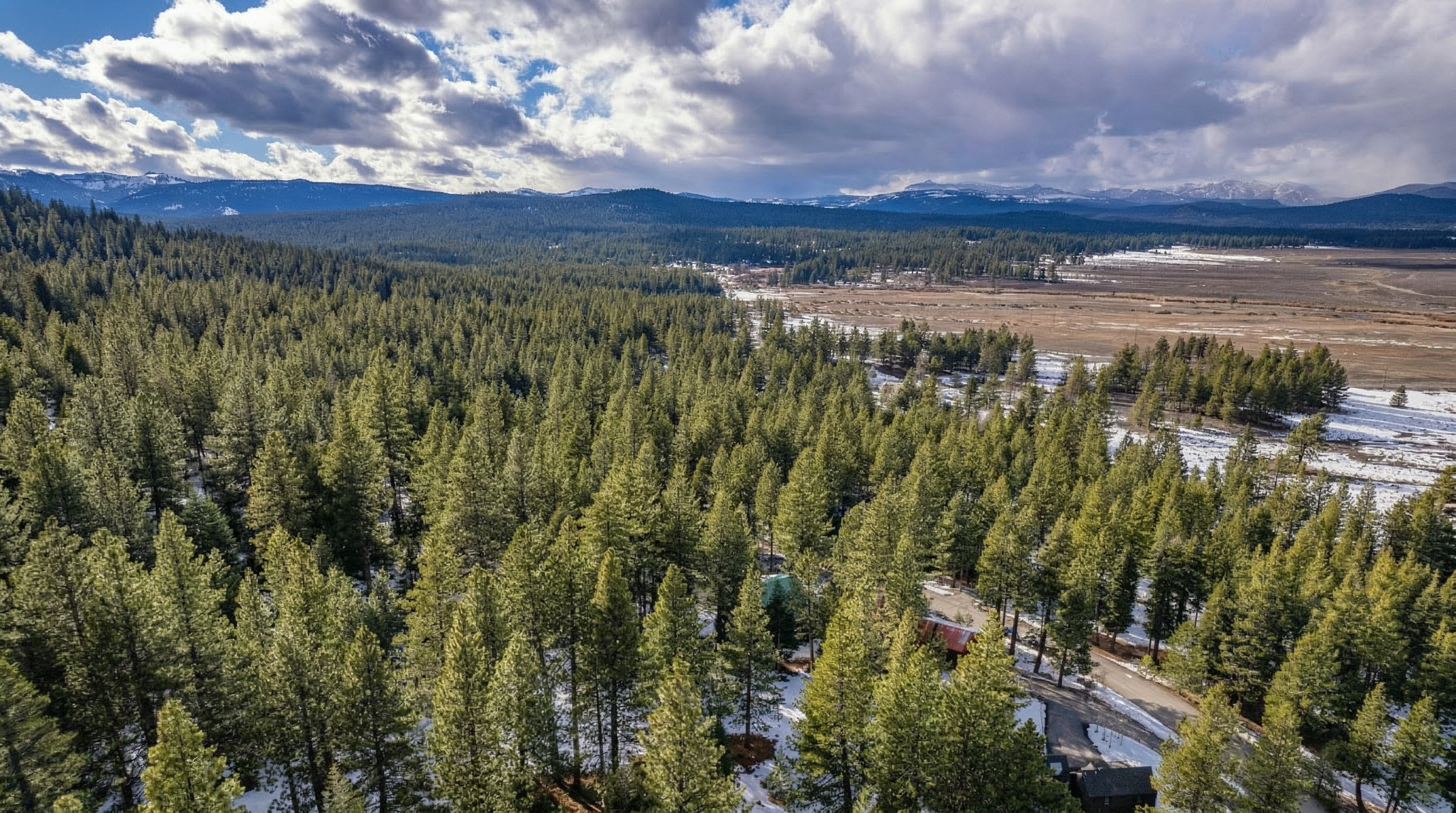 This is a scenic, high-altitude aerial perspective of a property nestled within a dense coniferous forest, showcasing the surrounding natural landscape. The image captures snow-dusted terrain, a mix of woodland and open meadow, and distant mountains under a dramatic partially cloudy sky. The location offers a secluded, tranquil setting ideal for those seeking proximity to nature and privacy.