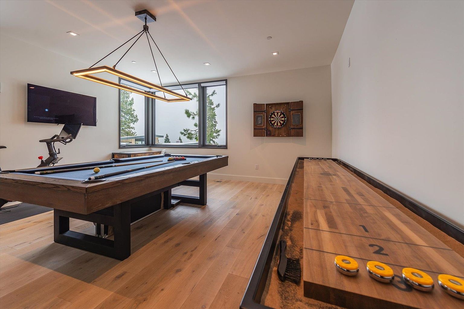 This interior shot showcases a recreational room featuring a pool table with cues and balls, a shuffleboard table with pucks, and a dartboard on the wall. A modern rectangular chandelier hangs above the pool table, and a television is mounted on the wall. The room has light hardwood flooring and neutral-colored walls, creating a relaxed and inviting atmosphere for entertainment and leisure.