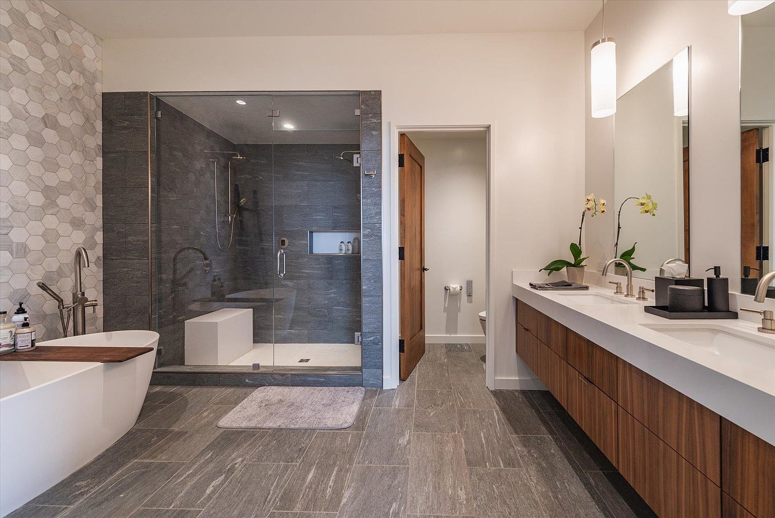 This is a modern primary bathroom featuring a freestanding bathtub, a glass-enclosed shower with gray tile, and a double vanity with a white countertop and wood cabinetry. The floor is covered in large gray tiles, and the walls are painted in a neutral tone. The overall impression is clean, luxurious, and well-designed.