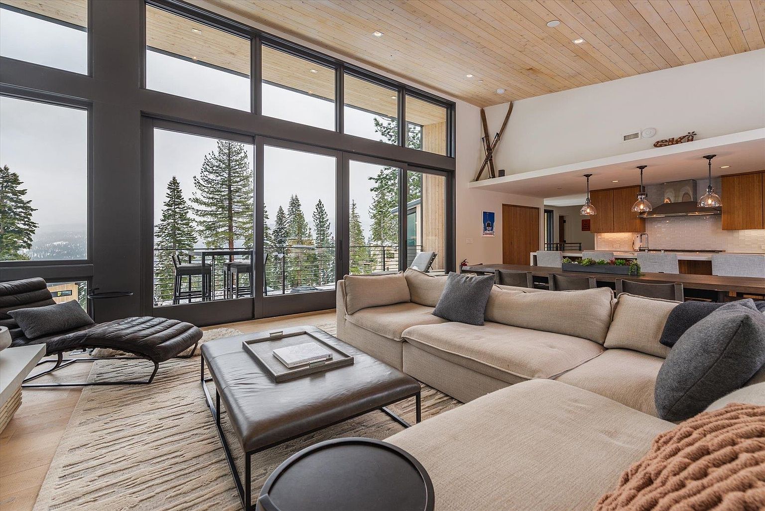 This is a spacious living room featuring a large sectional sofa, a leather ottoman, and a chaise lounge. The room has floor-to-ceiling windows that offer a view of the snow-covered trees outside. The open floor plan connects the living room to the kitchen and dining area, creating a seamless flow for entertaining.
