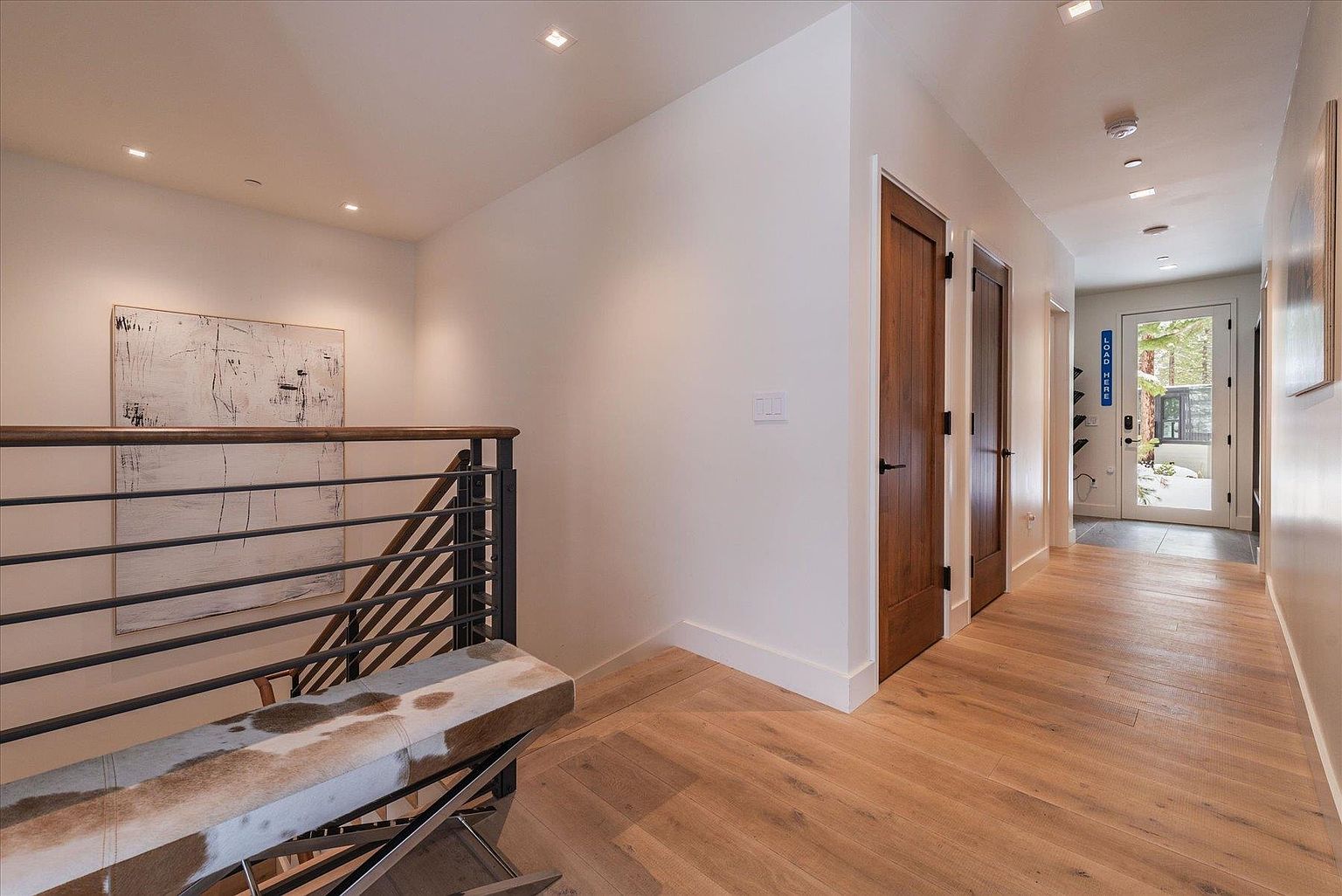 This interior shot showcases a well-lit hallway with hardwood flooring and white walls, leading to a bright entryway. A modern staircase with metal railings and a unique bench adds a stylish touch to the space. The overall impression is clean, contemporary, and inviting, highlighting the home's modern design elements.