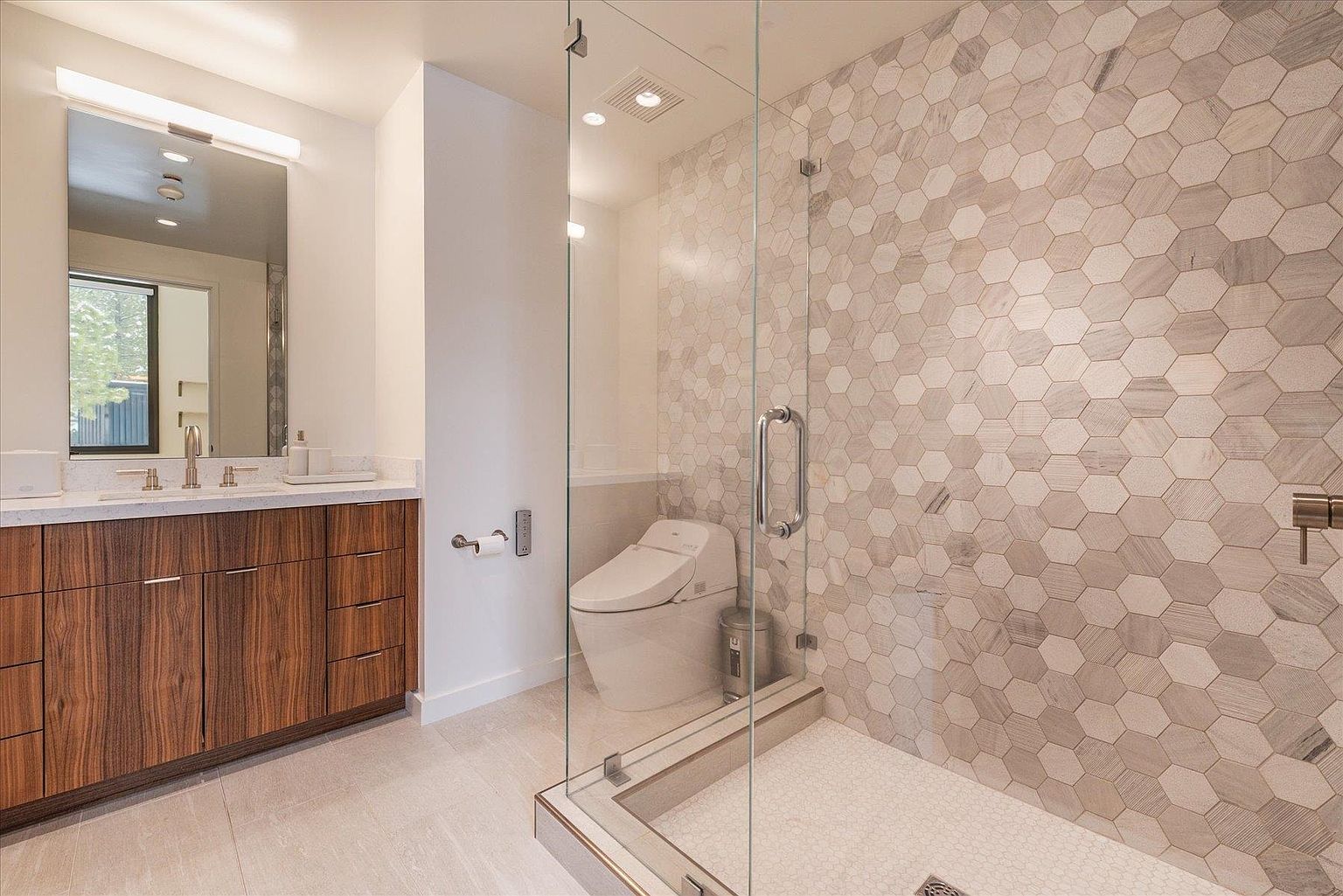 This is a modern primary bathroom featuring a glass-enclosed shower with hexagon tile walls and a white tile floor. A sleek wooden vanity with a white countertop and modern fixtures is visible, along with a toilet in the shower area. The overall impression is clean, contemporary, and luxurious.