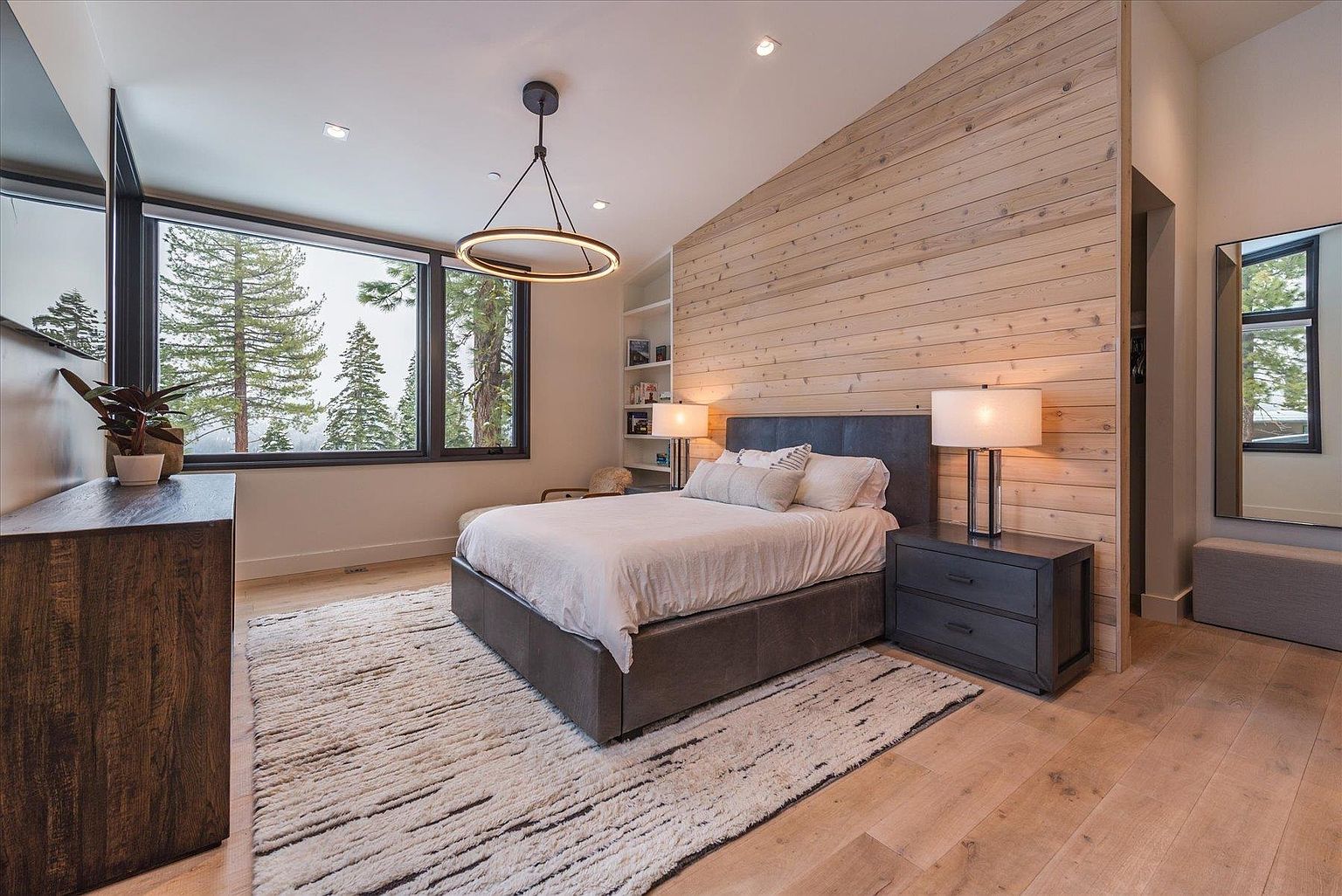 This is a primary bedroom featuring a modern design with a wood-paneled accent wall and a large window offering a view of the trees outside. The room includes a bed with a leather frame, a nightstand with a lamp, and a dresser. The overall impression is one of warmth and sophistication.
