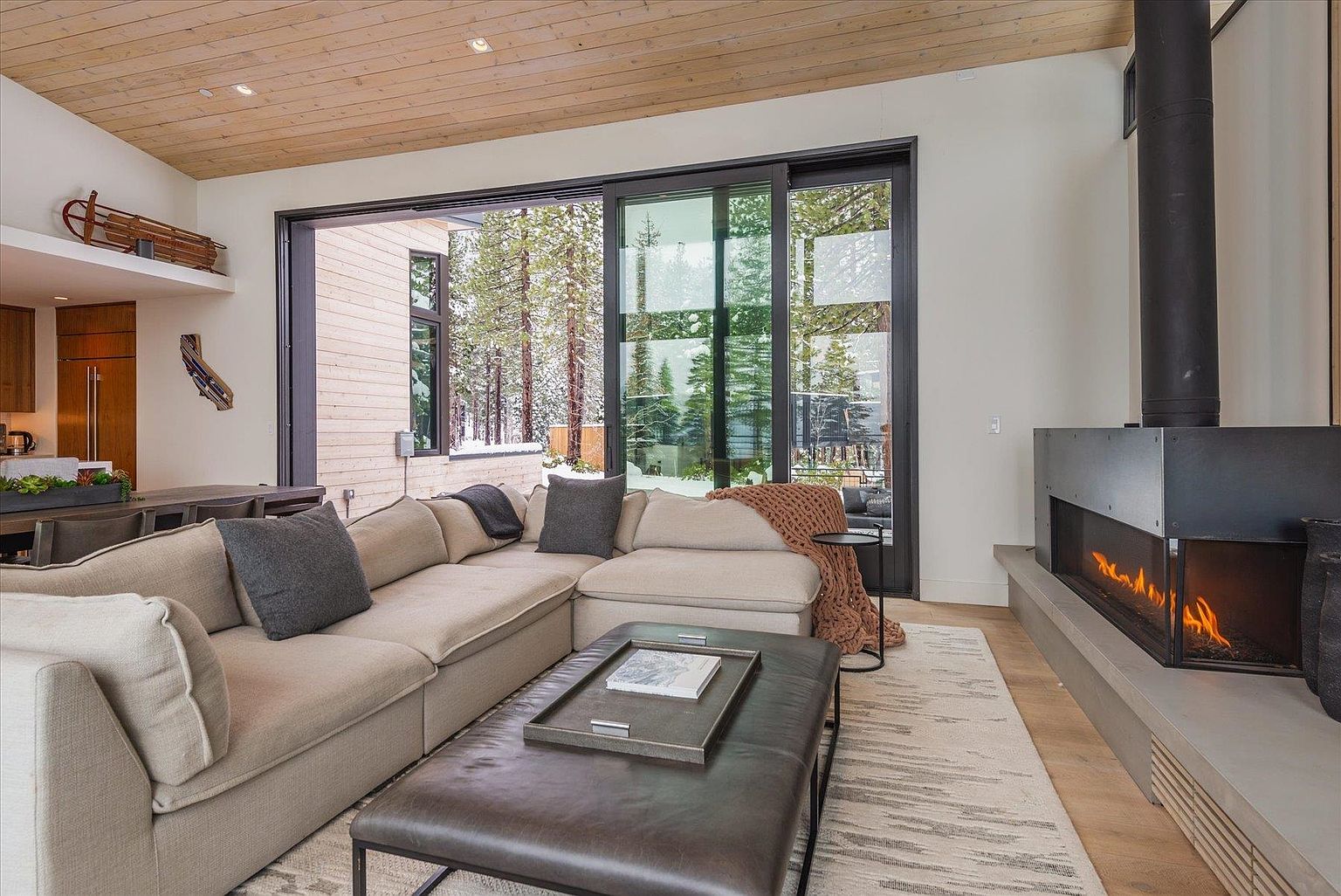 This is an interior shot of a modern living room featuring a large sectional sofa, a leather ottoman, and a contemporary fireplace. A large sliding glass door offers a view of a snowy outdoor scene, bringing natural light into the space. The room is decorated in neutral tones with wood accents, creating a warm and inviting atmosphere.