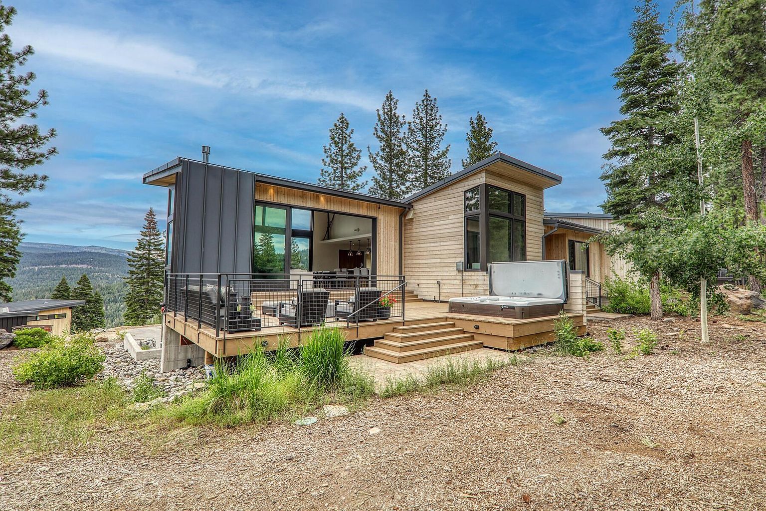 This image showcases a modern home's exterior, focusing on the deck and patio area. The deck features contemporary railings and outdoor seating, while a hot tub is situated on a separate platform accessible by steps. The home's design incorporates wood and metal elements, blending seamlessly with the natural surroundings.