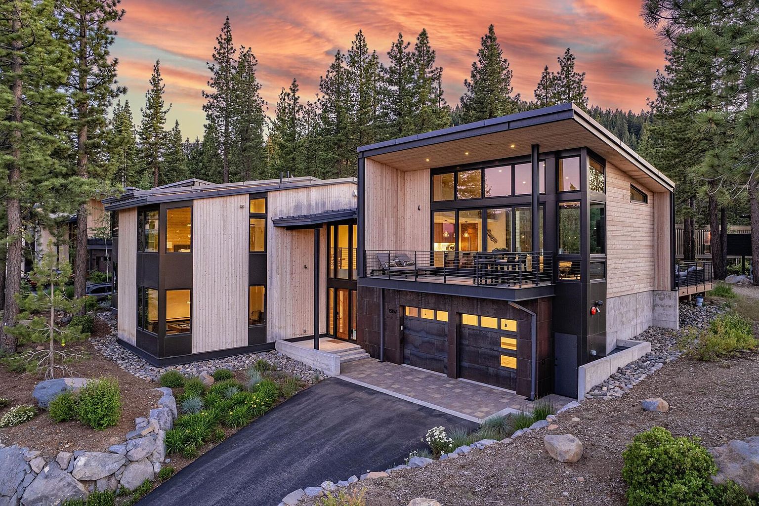This is a striking front view of a modern home with a unique architectural design. The exterior features a combination of light wood siding, dark metal accents, and large windows that offer ample natural light. A two-car garage is integrated into the design, and the landscaping includes a mix of rocks, plants, and a paved driveway, creating a sophisticated and inviting curb appeal.