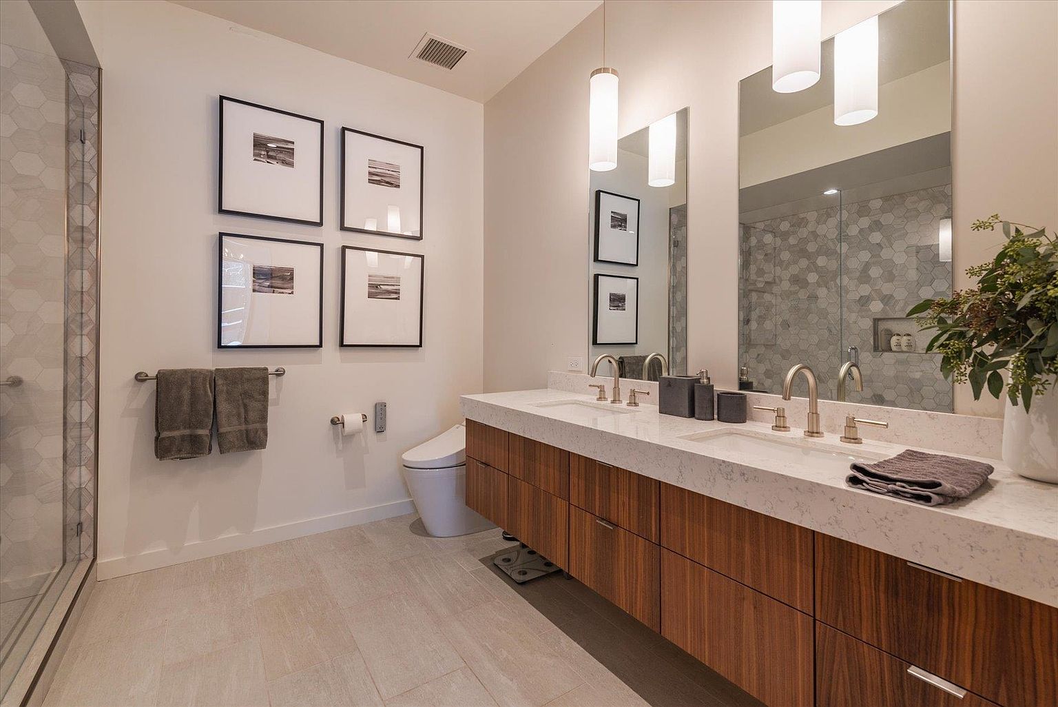 This is a well-lit primary bathroom featuring a double vanity with a white countertop and wood-grain cabinets. Above the vanity are two large mirrors with pendant lights hanging in front of them. The bathroom also includes a toilet, towel rack with towels, and a glass-enclosed shower with patterned tiles, creating a modern and luxurious feel.