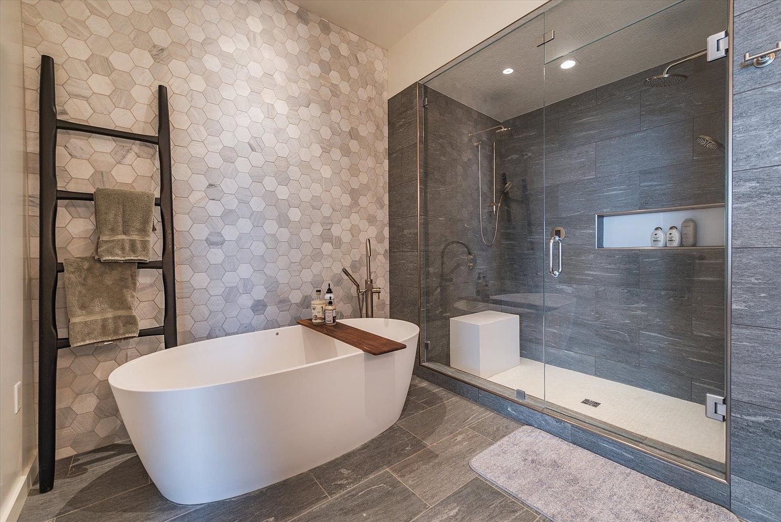 This is a luxurious primary bathroom featuring a freestanding white bathtub with a wooden tray, complemented by a decorative ladder with towels. The shower area is enclosed with glass and showcases gray tiled walls and a built-in niche. The flooring consists of large gray tiles, enhancing the spa-like atmosphere.