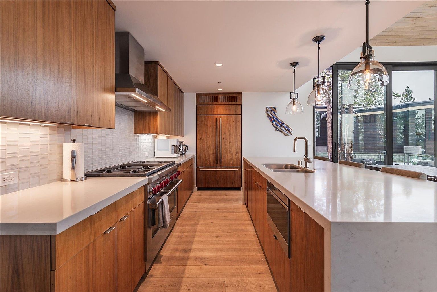 This is a well-lit, modern kitchen featuring wooden cabinetry and a long island with a white countertop. Stainless steel appliances are integrated throughout, and pendant lights hang above the island. The kitchen has a clean and contemporary design with a view to the outside.