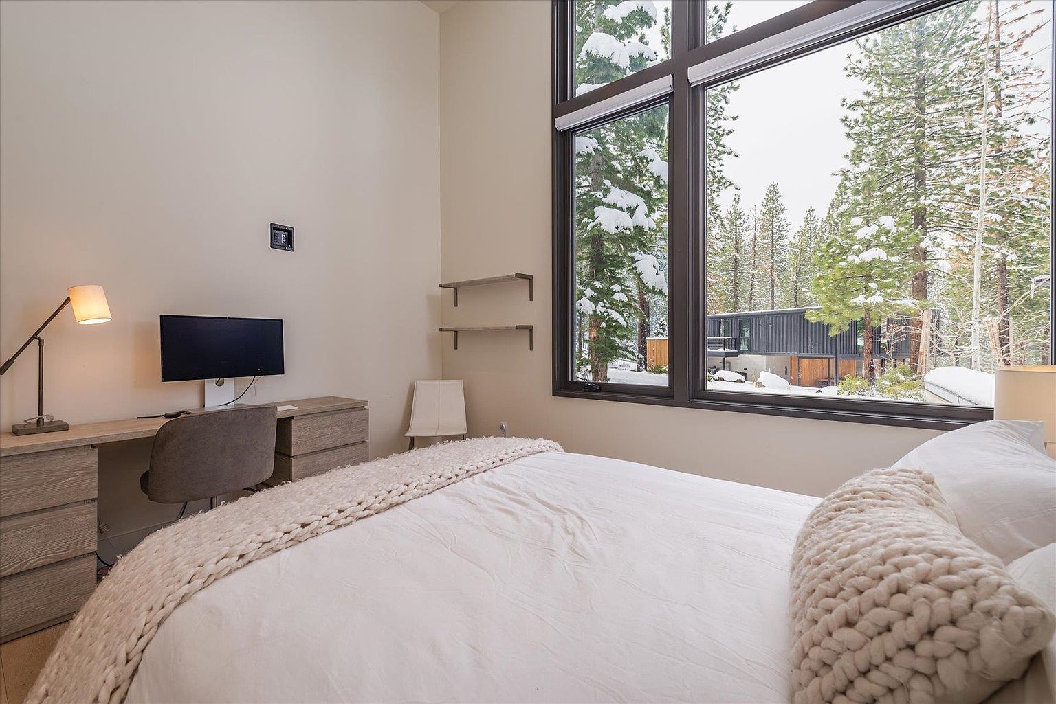 This bedroom features a modern aesthetic with a large window offering a view of snow-covered trees. The room includes a desk with a computer, a comfortable bed with a knitted throw, and minimalist shelving. The color palette is neutral, creating a serene and inviting atmosphere.