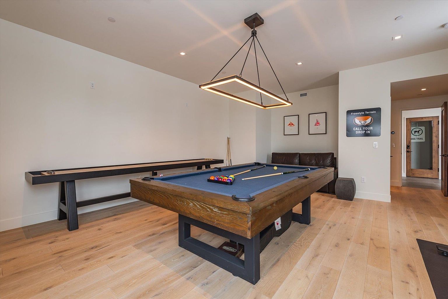This is an interior shot of a basement featuring a pool table with blue felt, a shuffleboard table, and a small seating area with a leather couch. The room is well-lit with recessed lighting and a modern rectangular chandelier above the pool table. The flooring is light-colored hardwood, and the walls are painted in a neutral tone, creating a recreational space.