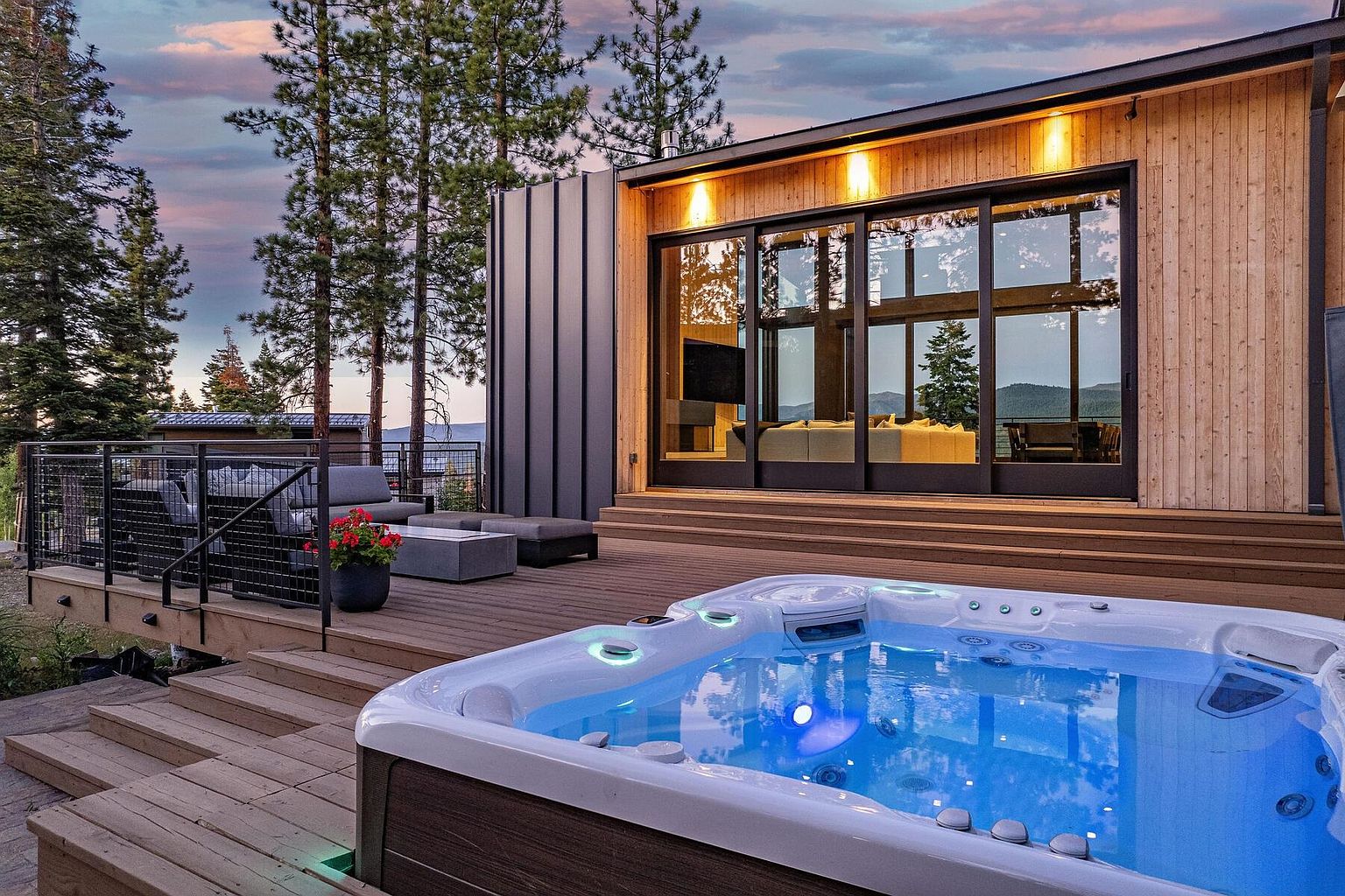 This image showcases a modern home's outdoor living space, featuring a spacious wooden deck with a hot tub as the focal point. The deck also includes comfortable seating arrangements and a sleek railing system, all set against a backdrop of tall trees and a picturesque sky. Large sliding glass doors connect the indoor and outdoor spaces, creating a seamless transition.