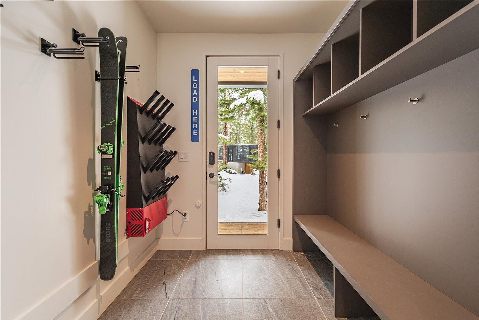 This interior shot showcases a modern entryway or hallway, featuring a ski rack and boot dryer on the left wall, suggesting a ski resort property. A gray built-in bench and shelving unit are on the right, providing ample storage. The door leads to a snowy outdoor scene, and the tile flooring adds a touch of elegance.
