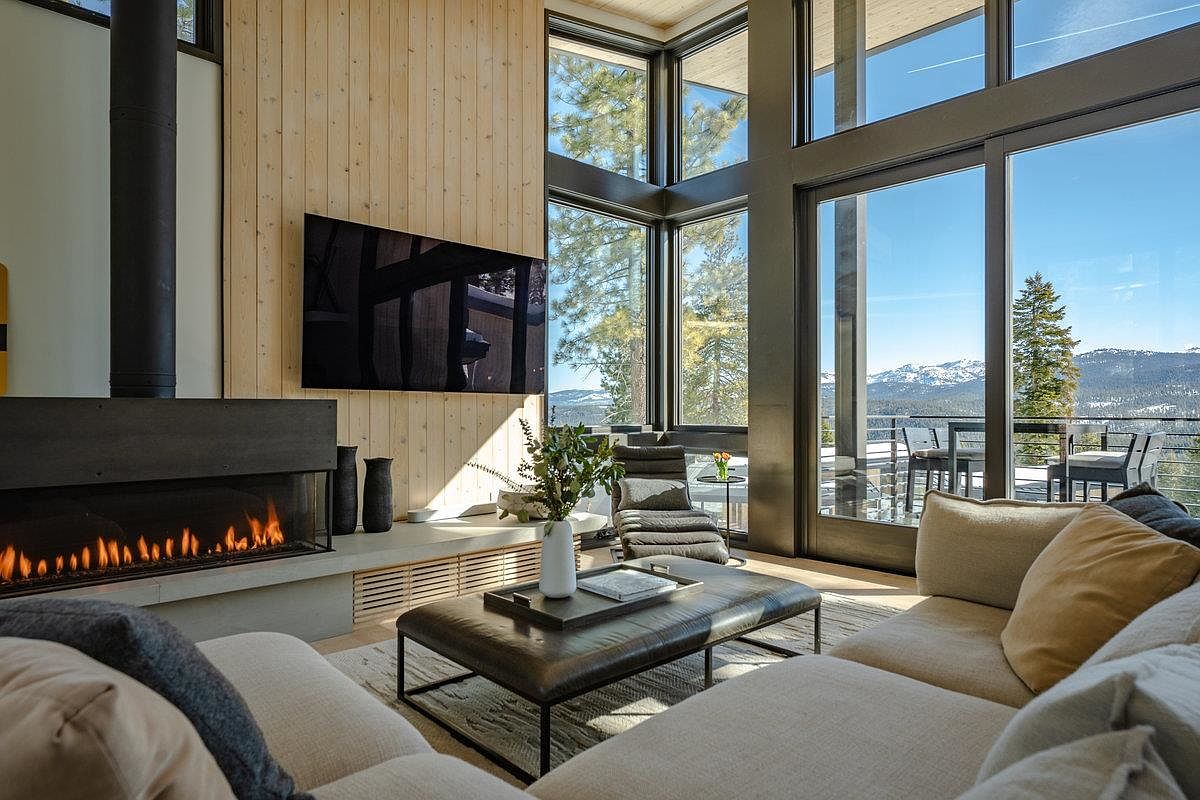 This is a luxurious living room featuring a modern fireplace, a large flat-screen TV mounted on a wooden accent wall, and floor-to-ceiling windows offering stunning mountain views. The room is furnished with a comfortable sectional sofa and a leather ottoman, creating a cozy yet sophisticated atmosphere. The open layout and natural light enhance the sense of spaciousness and elegance.