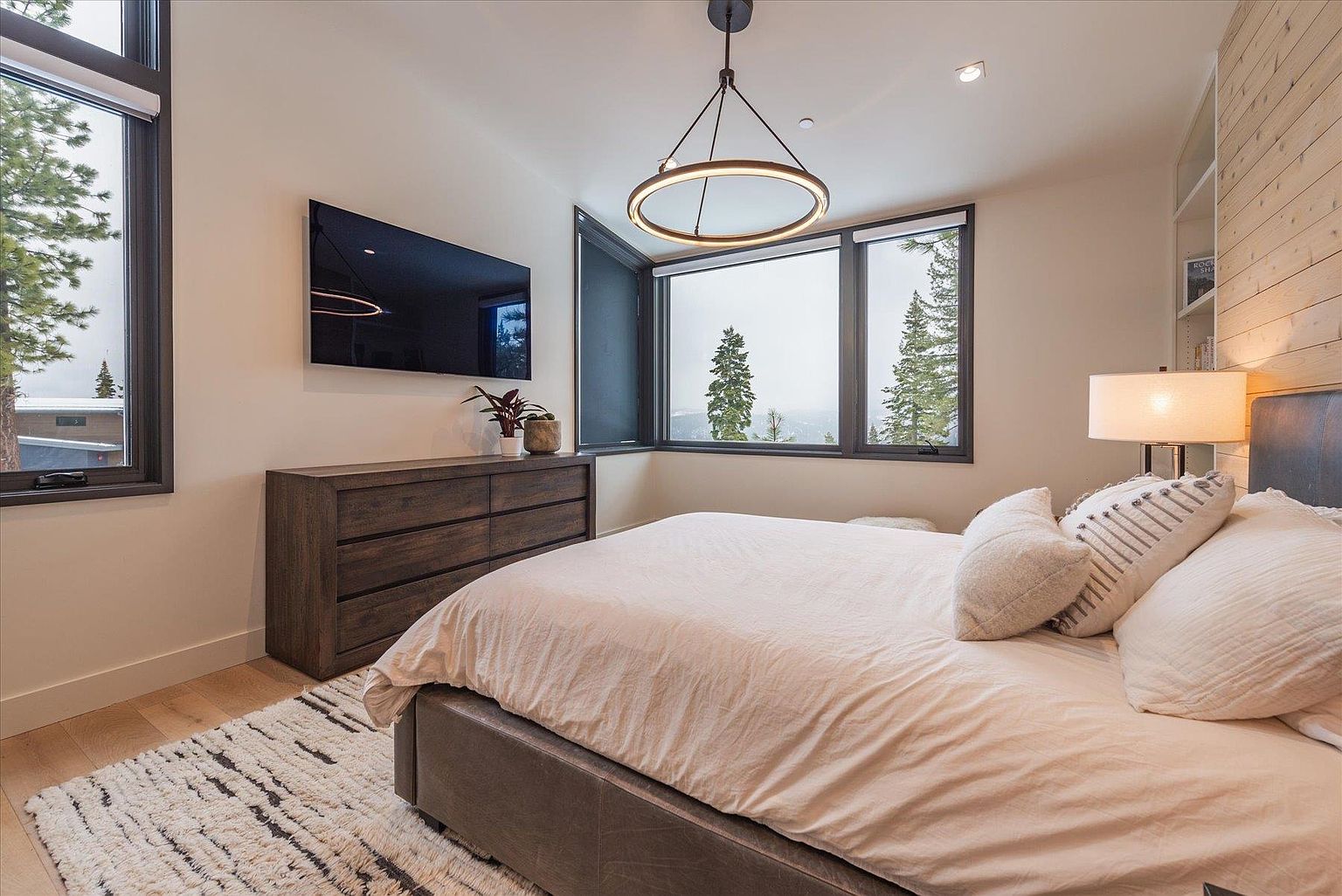 This is a primary bedroom featuring a modern design with a neutral color palette. The room includes a large bed with soft linens and decorative pillows, a wooden dresser with a flat-screen TV mounted above, and a large window offering a view of the outdoors. A stylish circular light fixture hangs from the ceiling, adding a touch of elegance.