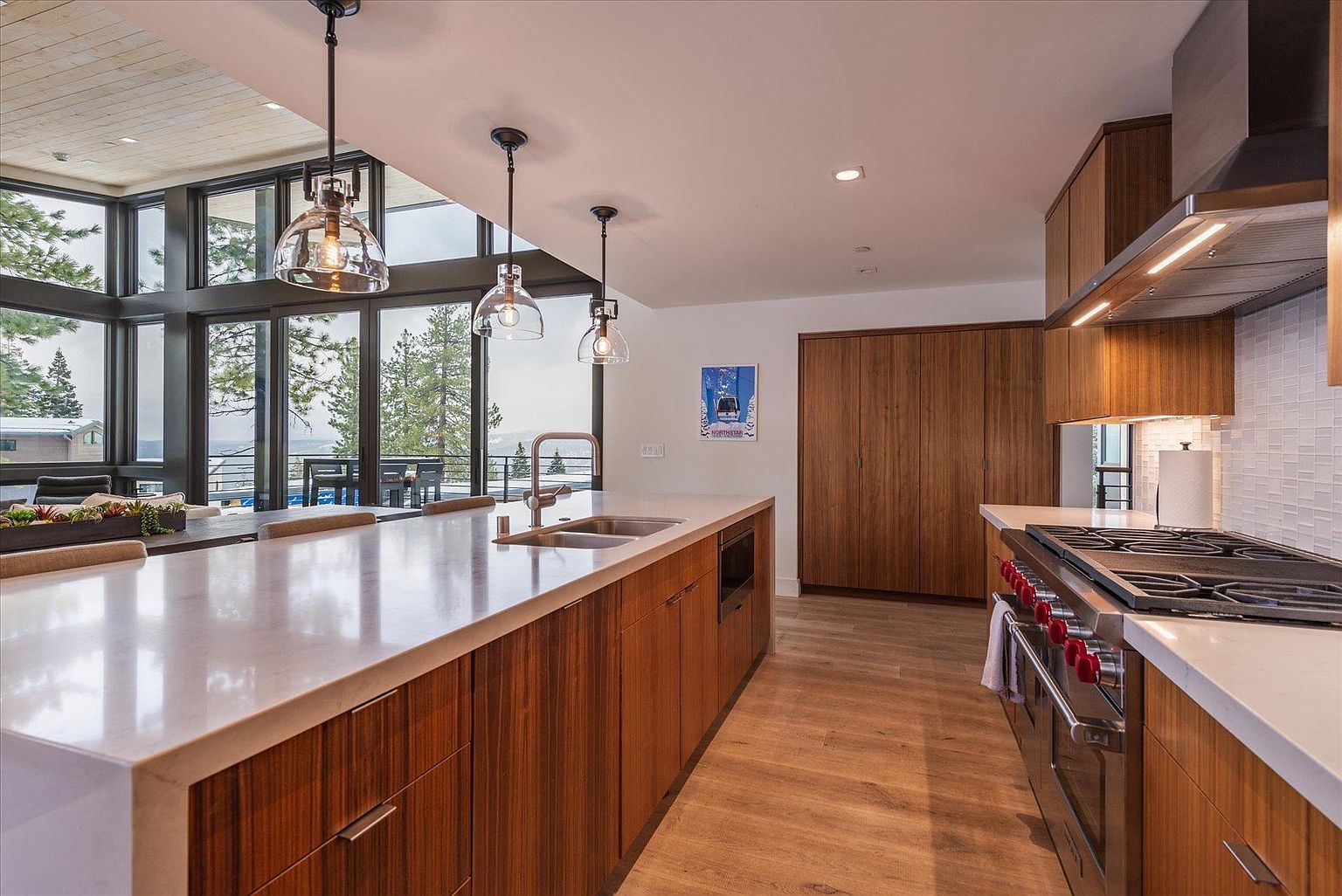 This is a bright and modern kitchen featuring wood cabinetry, a large island with a white countertop, and stainless steel appliances. Large windows offer a view of the outdoors, and pendant lights hang above the island. The kitchen has a clean and contemporary design with a focus on natural light and open space.