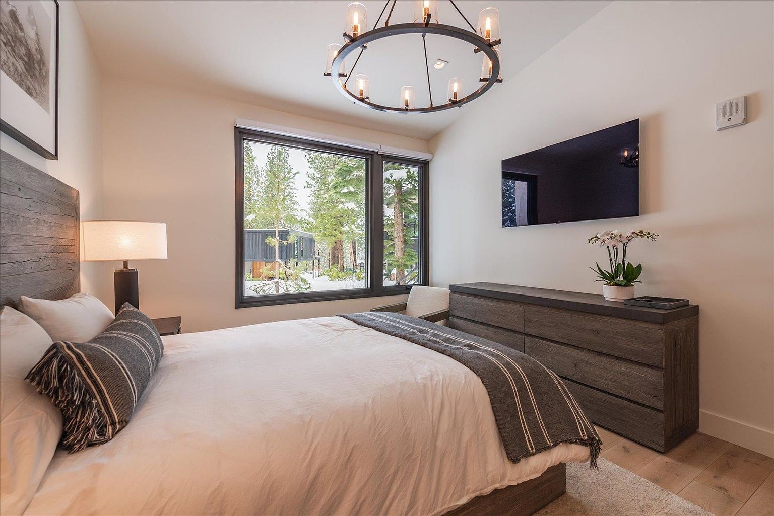 This is a well-lit primary bedroom featuring a king-size bed with a wooden headboard and a gray striped throw blanket. A modern chandelier hangs from the ceiling, and a large window offers a view of snow-covered trees. A dark wood dresser with a flat-screen TV mounted above it adds to the room's contemporary style.