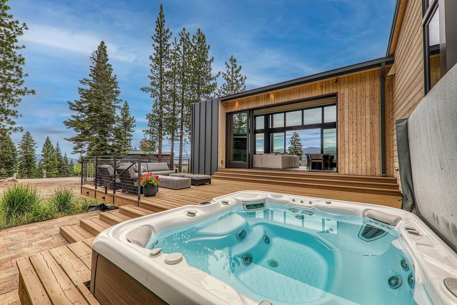 This image showcases a luxurious outdoor living space featuring a modern home with a spacious wooden deck. A hot tub takes center stage, inviting relaxation, while a comfortable seating area with a gray sofa and coffee table offers a place for socializing. The deck seamlessly connects to the interior through large sliding glass doors, and tall pine trees provide a scenic backdrop.