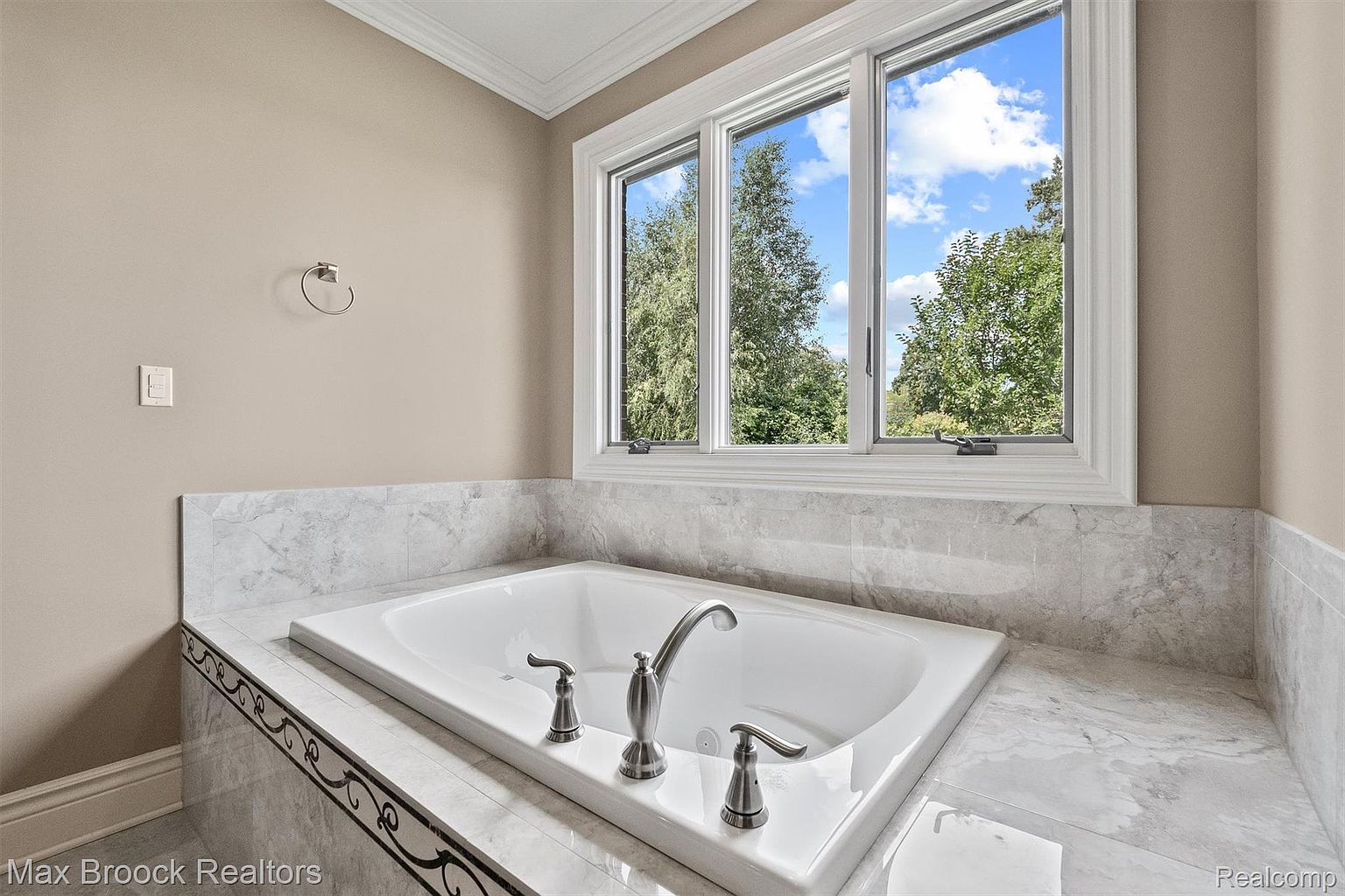 This image showcases a luxurious primary bathroom featuring a large, white soaking tub set against a backdrop of marble-like tile. A set of windows above the tub provides natural light and a view of the outdoors. The fixtures are brushed nickel, adding a touch of elegance to the space.