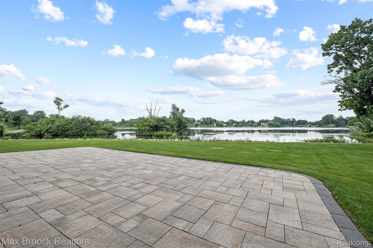 This image showcases a spacious patio area with a view of a serene lake. The patio is constructed with large, rectangular pavers, creating a modern and clean aesthetic. Lush green grass surrounds the patio, leading to the water's edge, enhancing the property's appeal for outdoor living and relaxation.