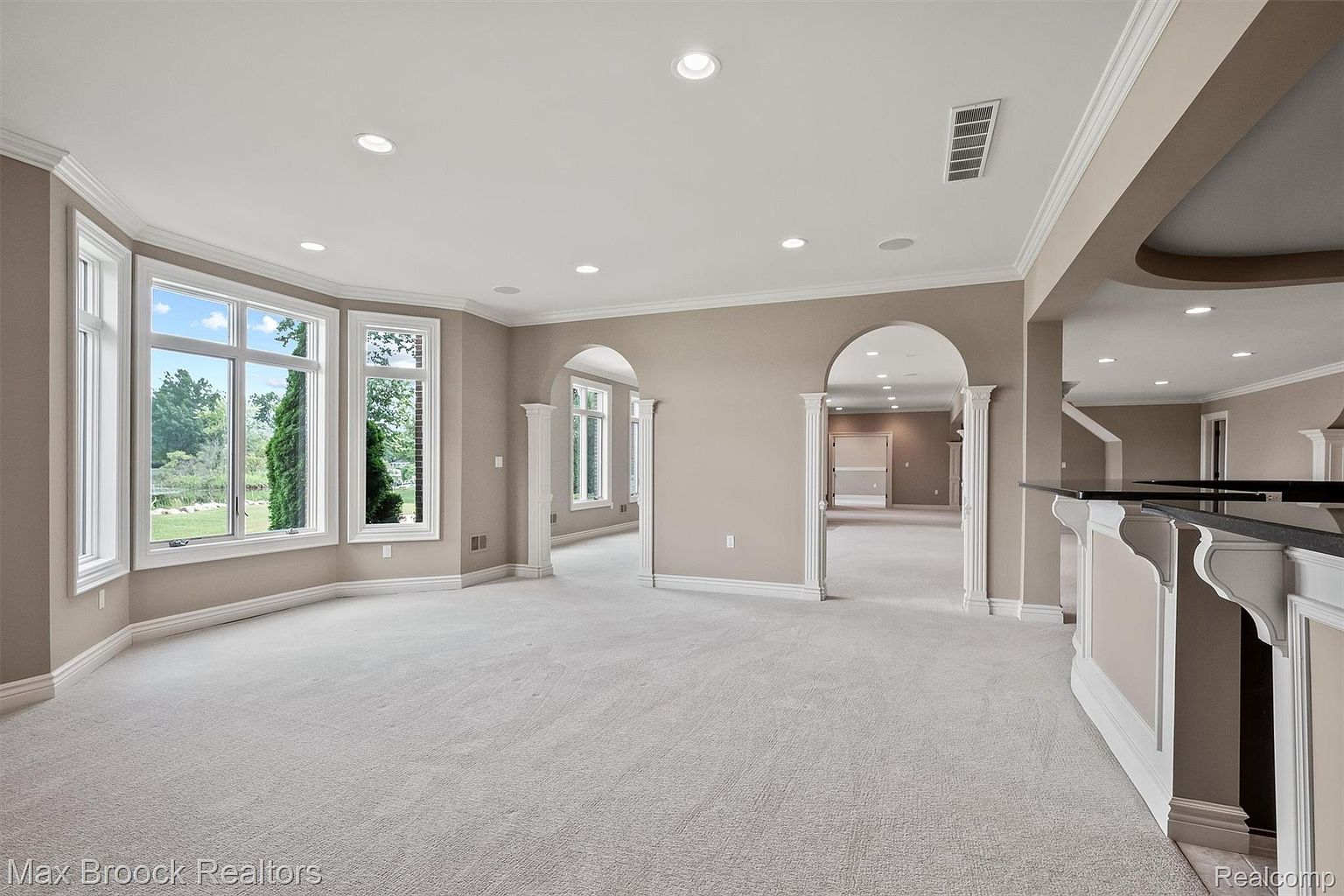 This is a spacious basement area featuring neutral-toned walls and light carpeting. The room is well-lit with recessed lighting and natural light from the bay windows. Arched doorways lead to other areas, and a bar area with a black countertop is visible on the right, suggesting a space designed for entertaining.