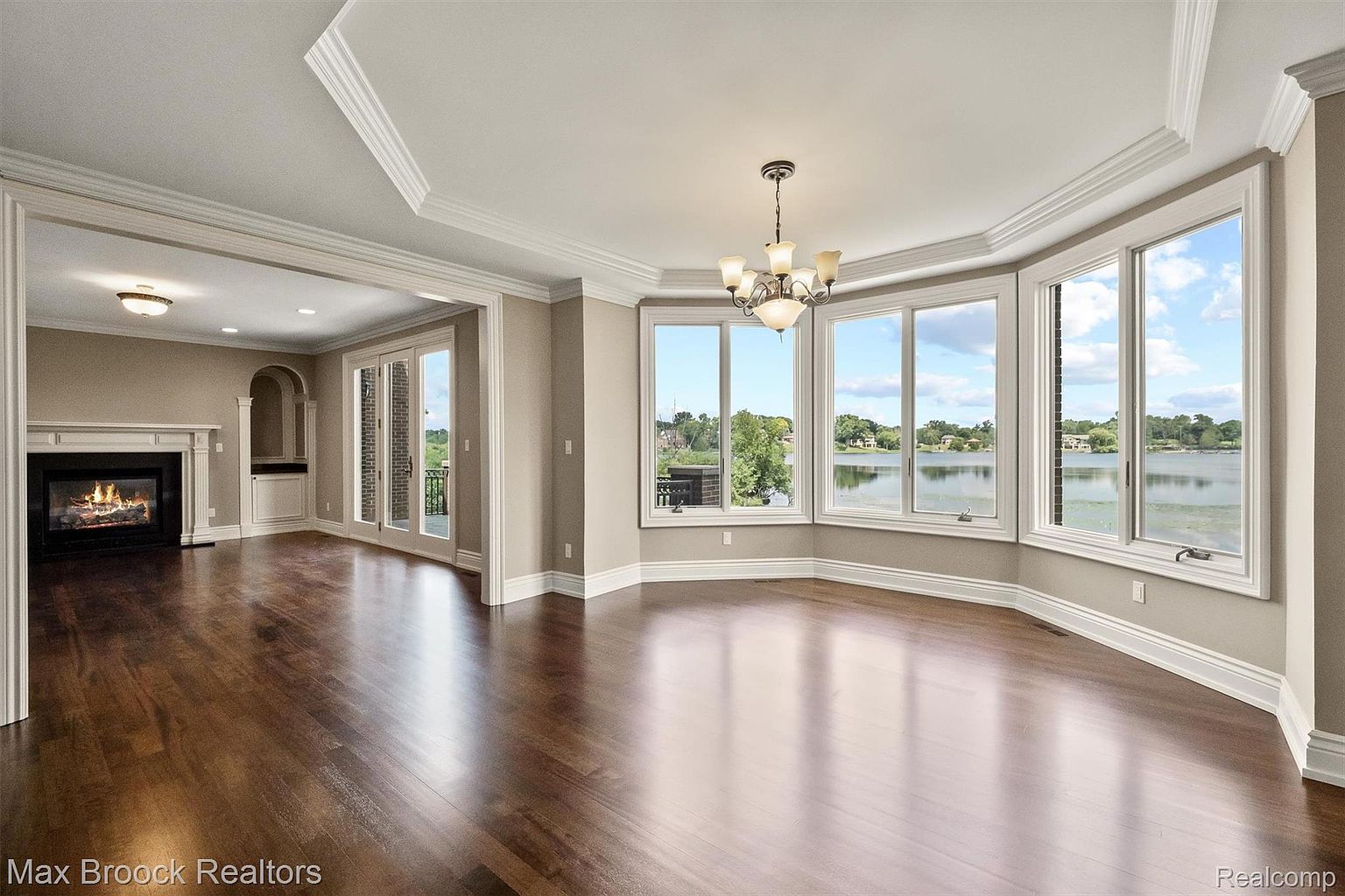 This is a bright and spacious dining room featuring dark hardwood floors and large bay windows offering a scenic view of a lake. The room is elegantly appointed with a chandelier and detailed crown molding, creating a luxurious and inviting atmosphere. A fireplace is visible in an adjacent room, adding warmth and character to the space.