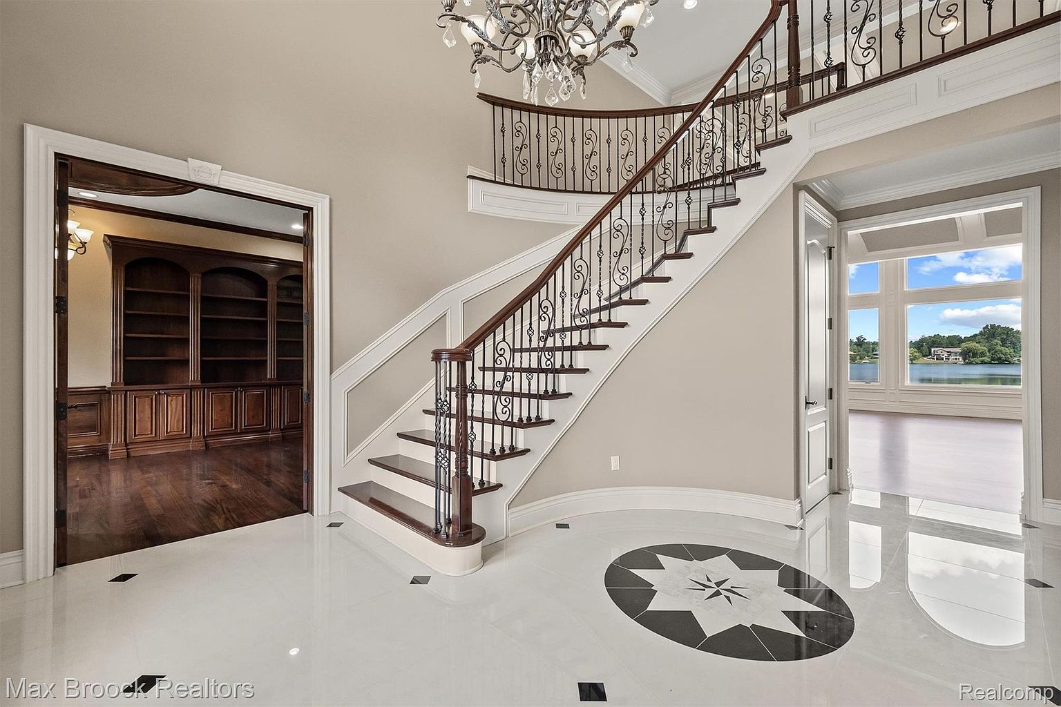 This grand foyer features a curved staircase with wrought iron railings, a striking black and white tile design on the floor, and a view into a wood-paneled office and a bright living area with lake views. The space is illuminated by a chandelier, creating an elegant and welcoming atmosphere, perfect for showcasing the home's luxurious appeal.