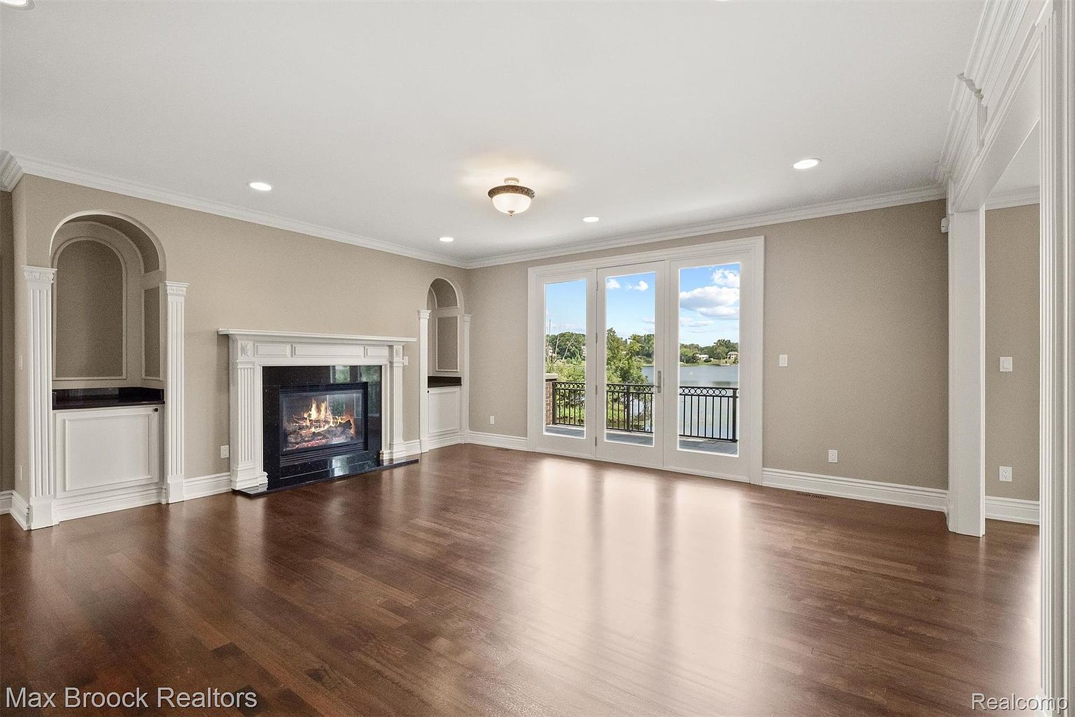 This is an interior shot of a luxurious living room featuring a dark hardwood floor, a fireplace with a black marble surround and white mantle, and built-in shelving units on either side. The room is well-lit with recessed lighting and natural light coming through the French doors that lead to a balcony overlooking a lake. The walls are painted in a neutral beige tone, and the overall impression is one of elegance and comfort.