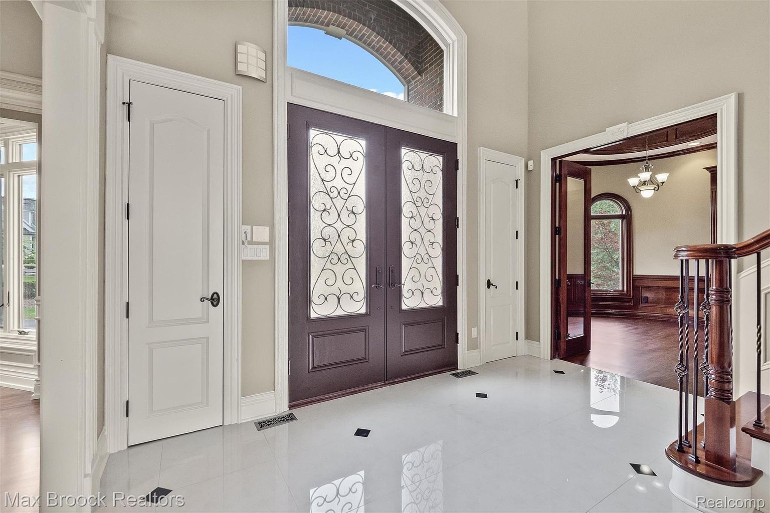 This interior shot showcases a grand foyer with a pristine white tile floor accented by black inlays. A set of ornate double doors serves as the main entrance, complemented by adjacent white doors and detailed trim work. The space leads into a room with rich wood paneling and a view through an arched window, creating an inviting and luxurious atmosphere.