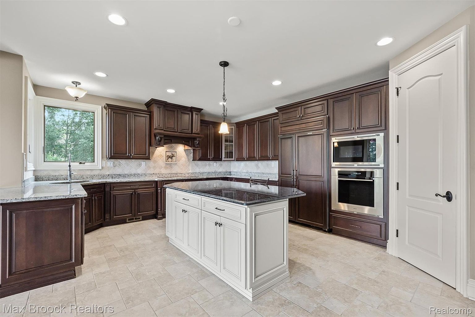 This is a well-lit kitchen featuring dark wood cabinetry, stainless steel appliances, and a contrasting white island with a dark countertop. The kitchen has a traditional style with a modern touch, and the tile flooring adds to the clean aesthetic. The perspective is a wide shot, showcasing the layout and features of the kitchen.