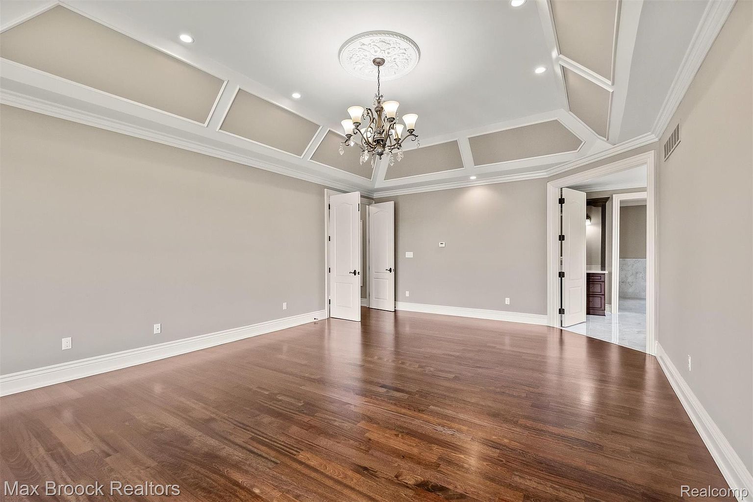 This is a spacious primary bedroom featuring hardwood floors, neutral-colored walls, and a tray ceiling with decorative molding and a chandelier. The room has multiple doorways, suggesting access to a bathroom or walk-in closet. The overall impression is one of elegance and comfort.