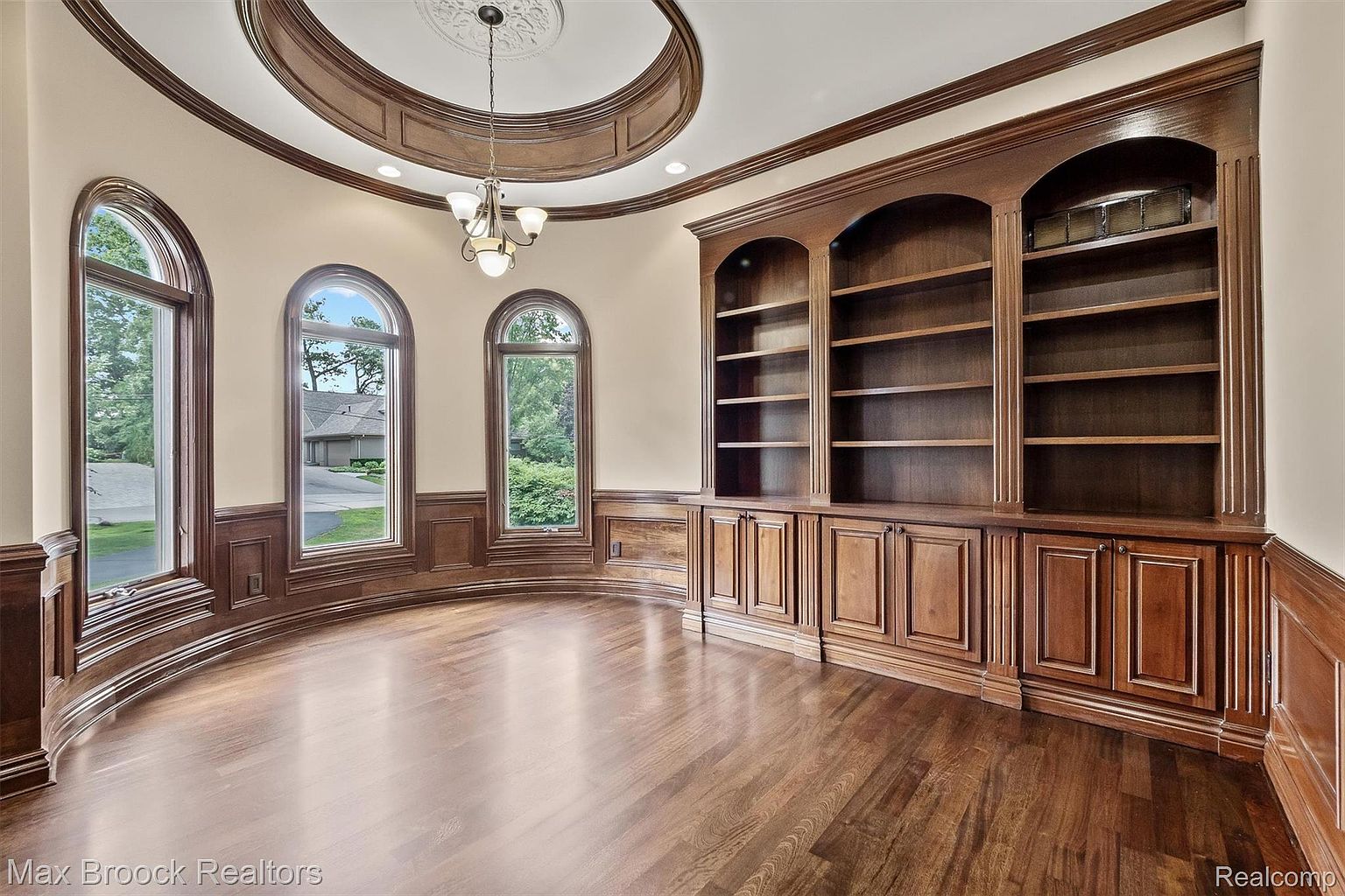 This is an interior shot of a luxurious office or study. The room features a curved wall with three arched windows, a large built-in wooden bookcase with cabinets, and rich hardwood flooring. The ceiling has an ornate circular design with a chandelier, creating an elegant and sophisticated atmosphere.