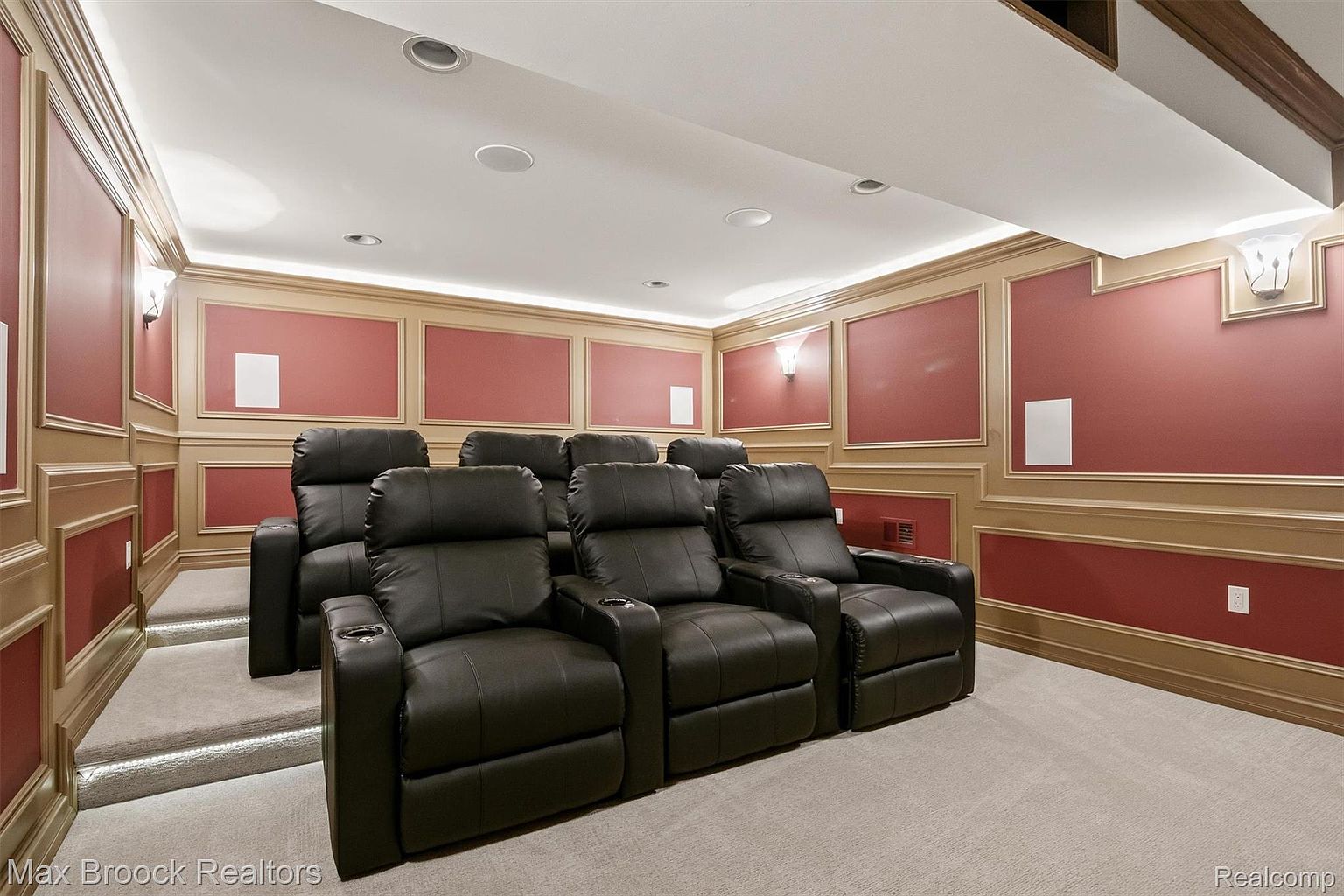This is an interior shot of a home theater. The room features tiered seating with black leather recliners, providing a comfortable viewing experience. The walls are adorned with decorative paneling, and recessed lighting illuminates the space, creating a cozy and immersive atmosphere.