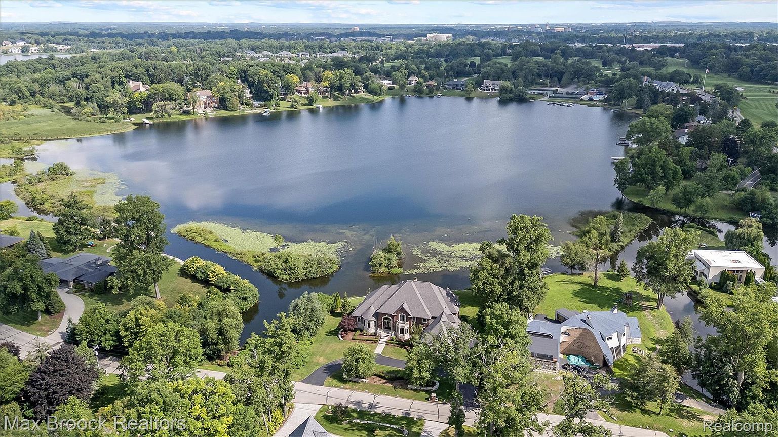 This aerial shot showcases a luxurious lakeside property with a sprawling house featuring a complex roofline and manicured grounds. The property is surrounded by mature trees and lush landscaping, offering privacy and a serene setting. The lake provides a stunning backdrop, enhancing the overall appeal of this high-end real estate offering.